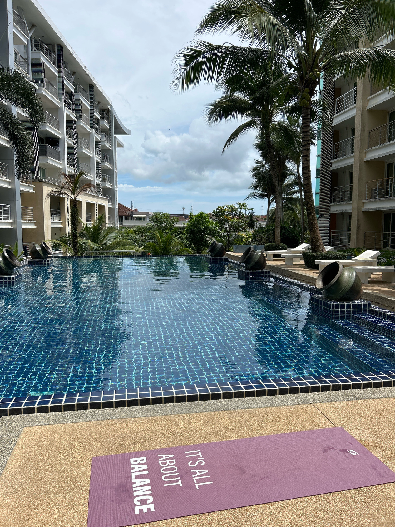The image depicts a swimming pool area at a resort in Kathu, Thailand. The pool is tiled in blue and surrounded by a beige patio. Large, dark green ceramic pots are placed along the edge of the pool. In the background, modern white buildings with balconies and palm trees can be seen. On the patio in the foreground, a purple yoga mat with white text that reads "IT'S ALL ABOUT BALANCE" is laid out. The sky is cloudy, suggesting an overcast day.