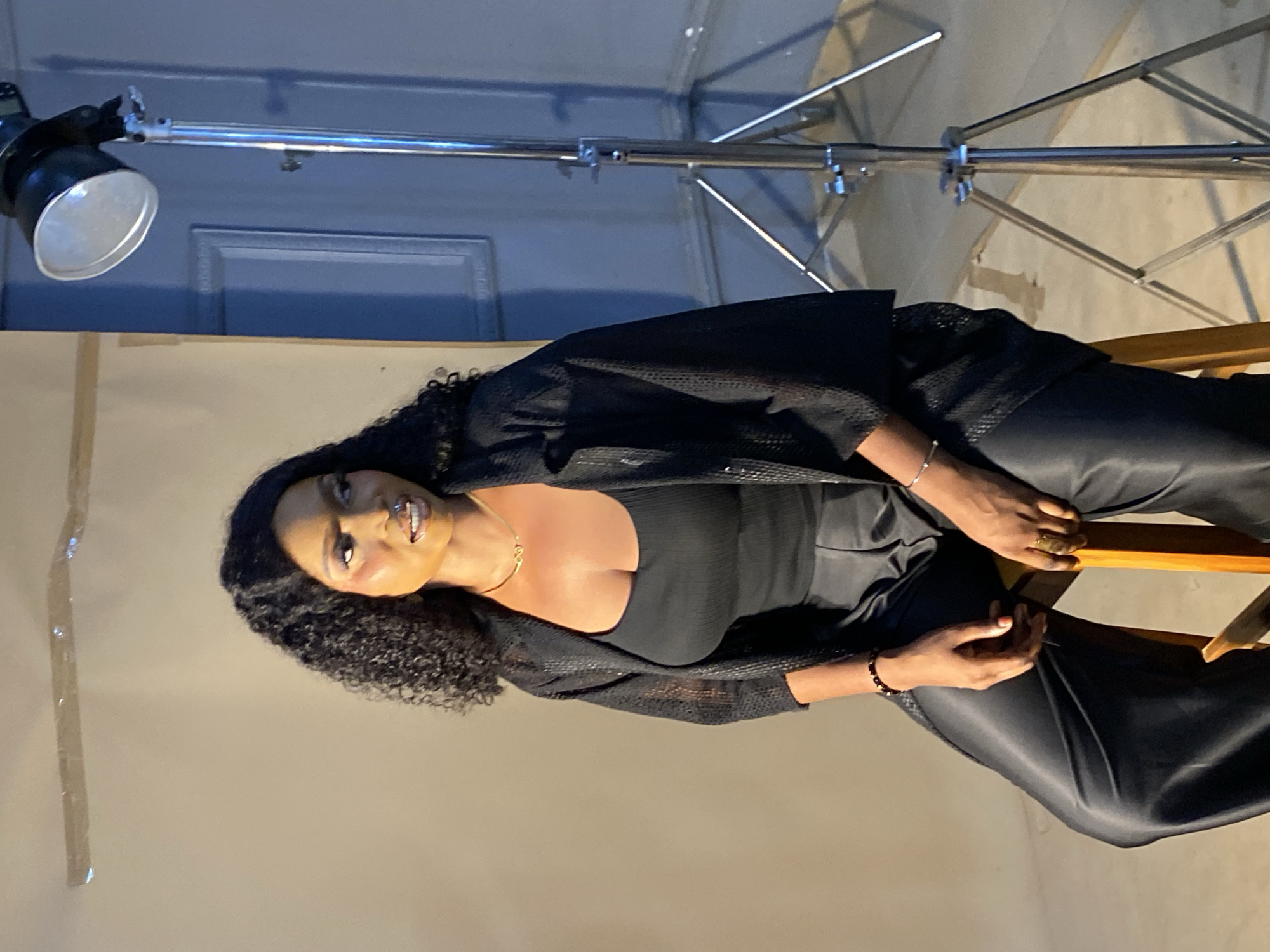 In a studio setting, a Black woman is seated on a wooden stool against a neutral backdrop. She has curly black hair and is wearing a black sleeveless top, a sheer black open-front jacket, and black satin pants. Her makeup is bold, with emphasis on her eyes and lips. She is adorned with a delicate gold necklace, beaded bracelets on her wrists, and a ring on her right hand. To her right, a professional studio light on a stand is positioned. The background transitions from a light beige on the left to a darker blue-grey on the right, with a framed element visible on the blue-grey wall. The floor appears to be concrete. The scene is indicative of a photoshoot. The prompt requests a specific location in Bukuru, Nigeria, but this information cannot be determined from the image itself.
