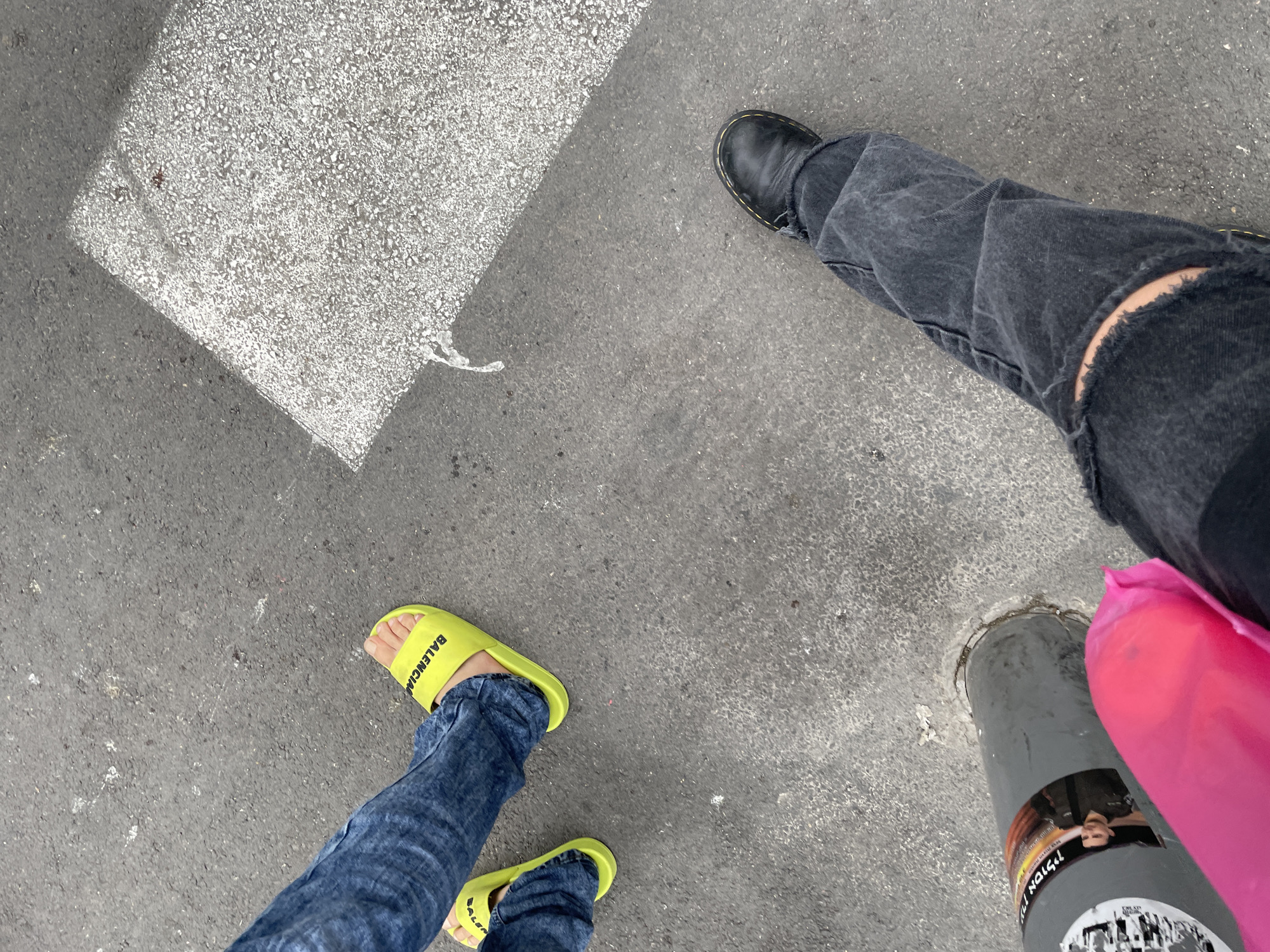 The scene shows a view from above, depicting portions of two individuals standing on a gray asphalt surface, likely a street or sidewalk. One person is wearing distressed black jeans and a black leather boot, while the other is wearing blue jeans with bright yellow slides marked "BALENCIAGA." A gray metal pole with affixed promotional stickers and a large, pink plastic bag sits near the person wearing jeans and a boot. Part of a light-colored concrete slab is visible in the upper-left portion of the frame. The scene is consistent with the urban environment of Tel Aviv, Israel.