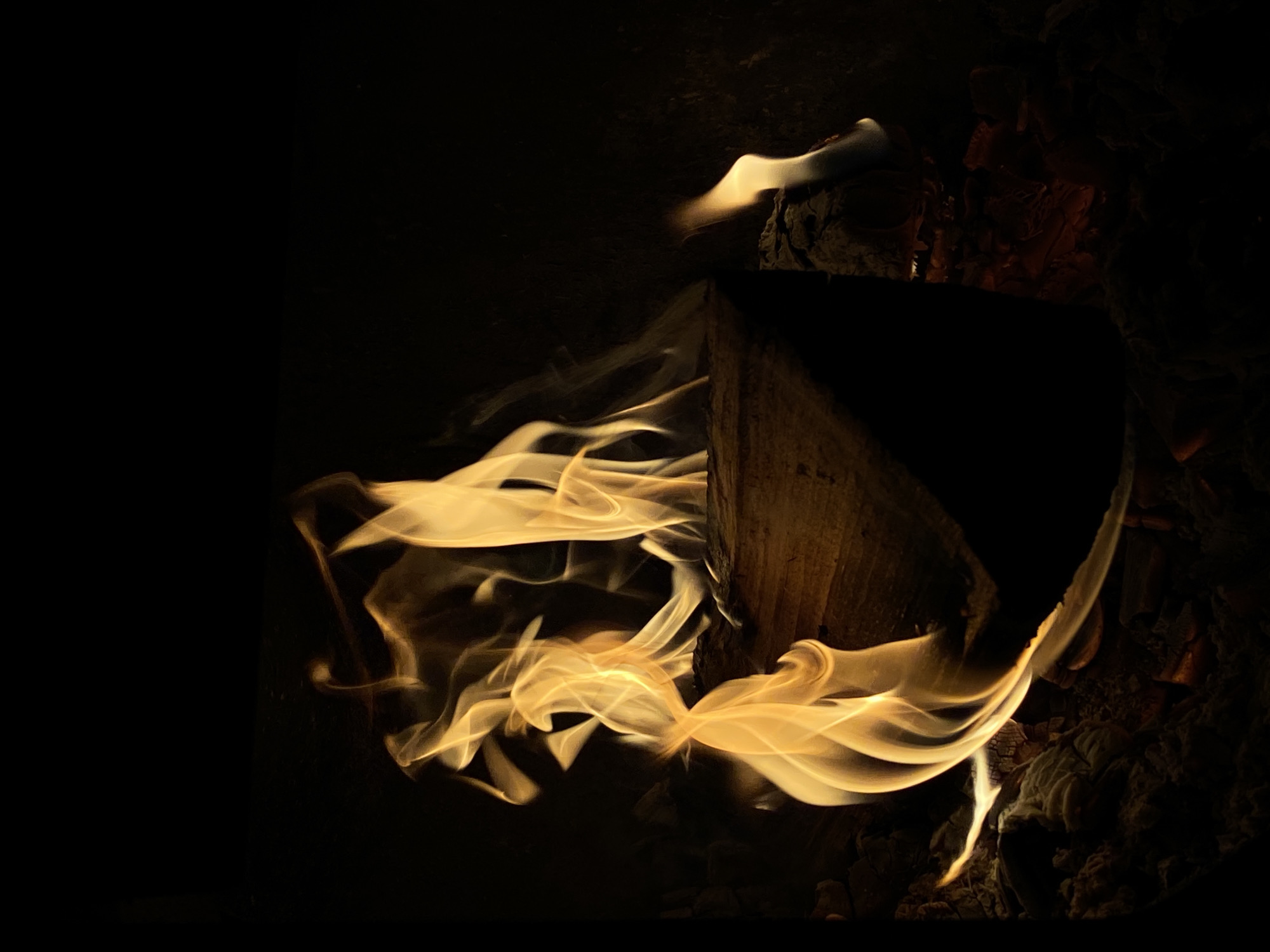 The image depicts a dark scene with a fire burning next to a piece of wood. The flames are flowing and swirling, illuminated against the black background. To the right of the wood is a pile of what appears to be charred material. It is not possible to ascertain anything about the location of this scene being in the city of Фоки, Russia from the visual information contained in this image.