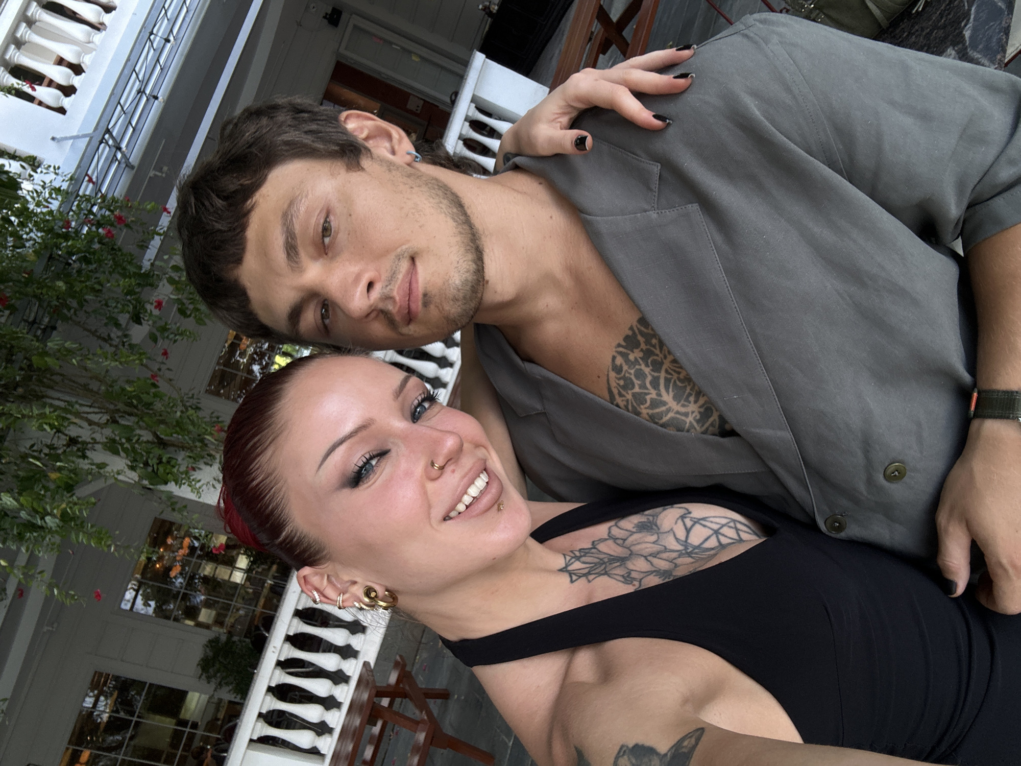 A medium shot captures a young couple taking a selfie outdoors in Hoi An, Vietnam. The woman, on the left, has red hair pulled back, blue eye makeup, and several piercings, including large hoop earrings and a nose ring. She wears a black halter top that reveals a large floral tattoo on her chest. She is smiling broadly, with her head tilted towards the man. The man, on the right, has short brown hair and a light beard. He wears a grey blazer over a shirt with a tattoo visible on his chest. His arm is around the woman, and her hand rests on his shoulder. They are positioned in front of a building with white ornate railings and balconies, with greenery hanging overhead. In the background, wooden tables and chairs are visible, suggesting an outdoor dining area. The overall lighting indicates it is daytime.
