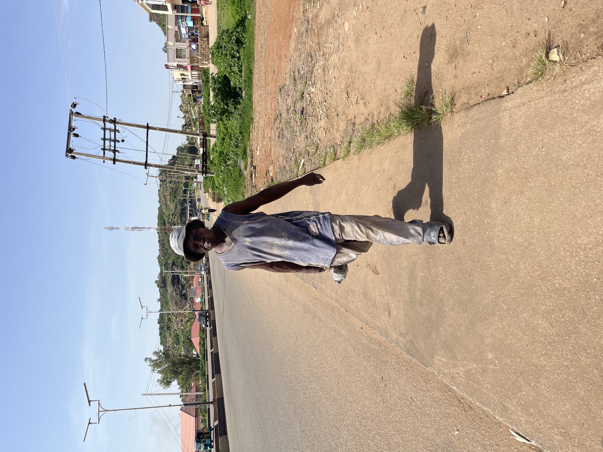 A man walks on a sidewalk in Gura Top, Nigeria. He is wearing a blue tank top, beige pants, and a grey bucket hat. He is carrying a grey grinder in his right hand. The sidewalk is made of concrete and has grass growing through cracks. A paved road runs alongside the sidewalk, with traffic and buildings in the background. The sky is clear and blue.