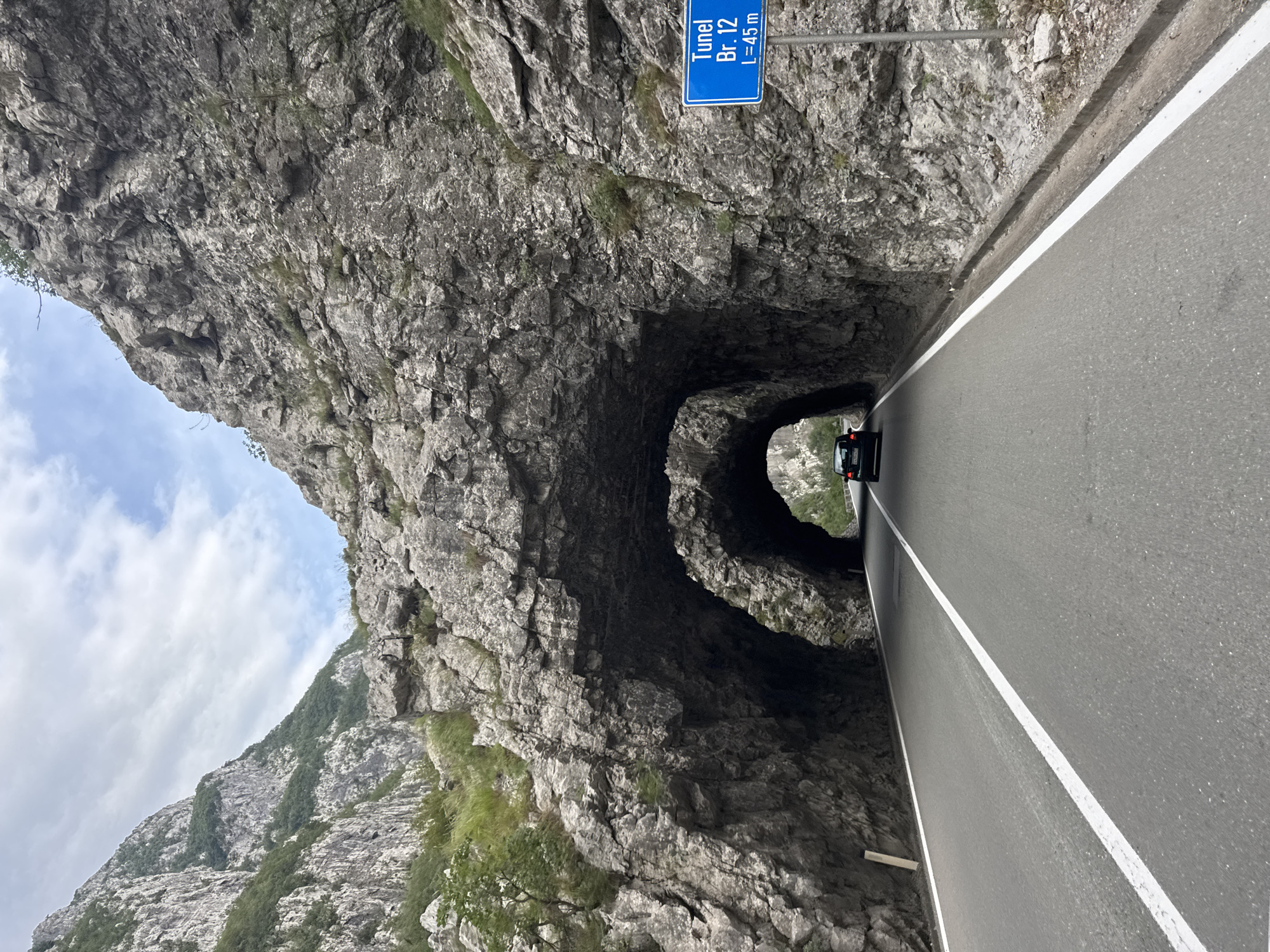 The image depicts a view from a car driving on a road through a mountainous area. The road is asphalt with a white line marking the lane. On the right side of the road, there is a blue sign that reads "Tunel Br. 12 L=45 m", indicating a tunnel numbered 12 with a length of 45 meters. The road leads to and passes through a large rock formation that has been carved into two arched tunnels. The tunnels are dark, and a black car is visible driving through the first tunnel. The surrounding environment is characterized by steep, rocky mountains with sparse vegetation. The sky is partly cloudy. The location is identified as Vlahovići, Montenegro.