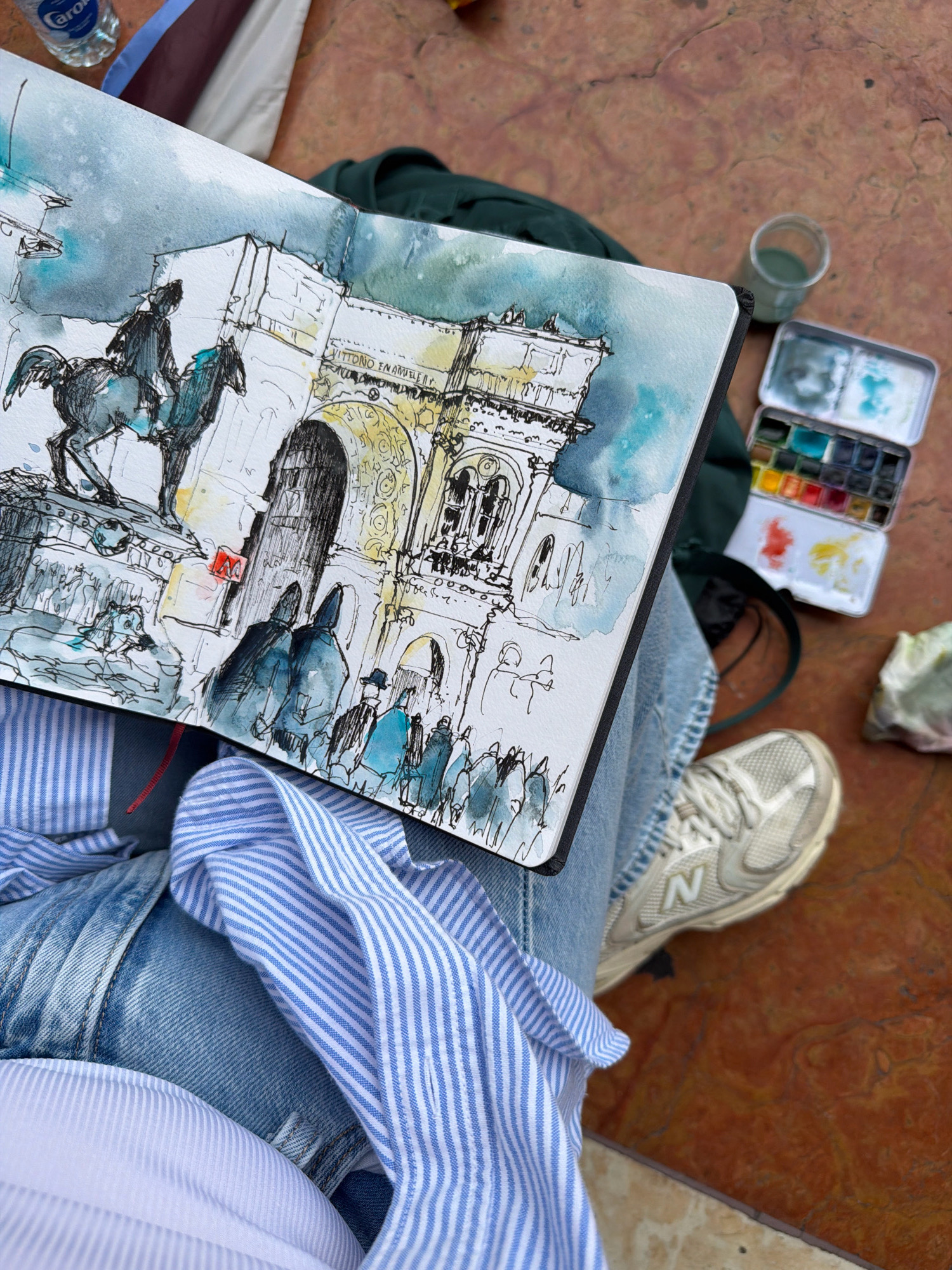 The image shows a person's lap with a sketchbook open, displaying a watercolor and ink sketch of the Arch of Constantine in Rome, not Milan. The sketch depicts a horse-mounted statue in the foreground, with the archway and figures in the background. Adjacent to the sketchbook is a metal watercolor tin filled with various colors, a glass jar with a greenish liquid, and a crumpled white tissue. The person is wearing light blue jeans and a blue and white striped shirt, with a white shirt underneath. A white sneaker with an "N" logo is visible on the person's right foot. The background consists of a reddish-brown textured surface, likely a table or pavement. A plastic water bottle is partially visible in the upper left corner.