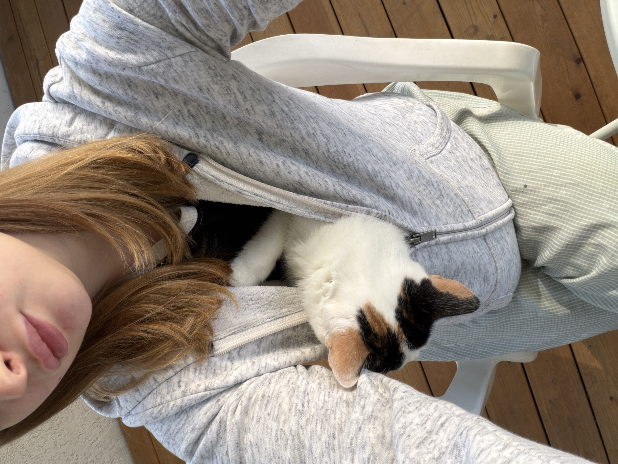 A light-skinned person with light brown hair is sitting in a white plastic chair. The person is wearing a gray hooded sweatshirt. Inside the partially unzipped sweatshirt is a calico cat. The background is a wooden deck. The location is Rossiyskiy, Russia.