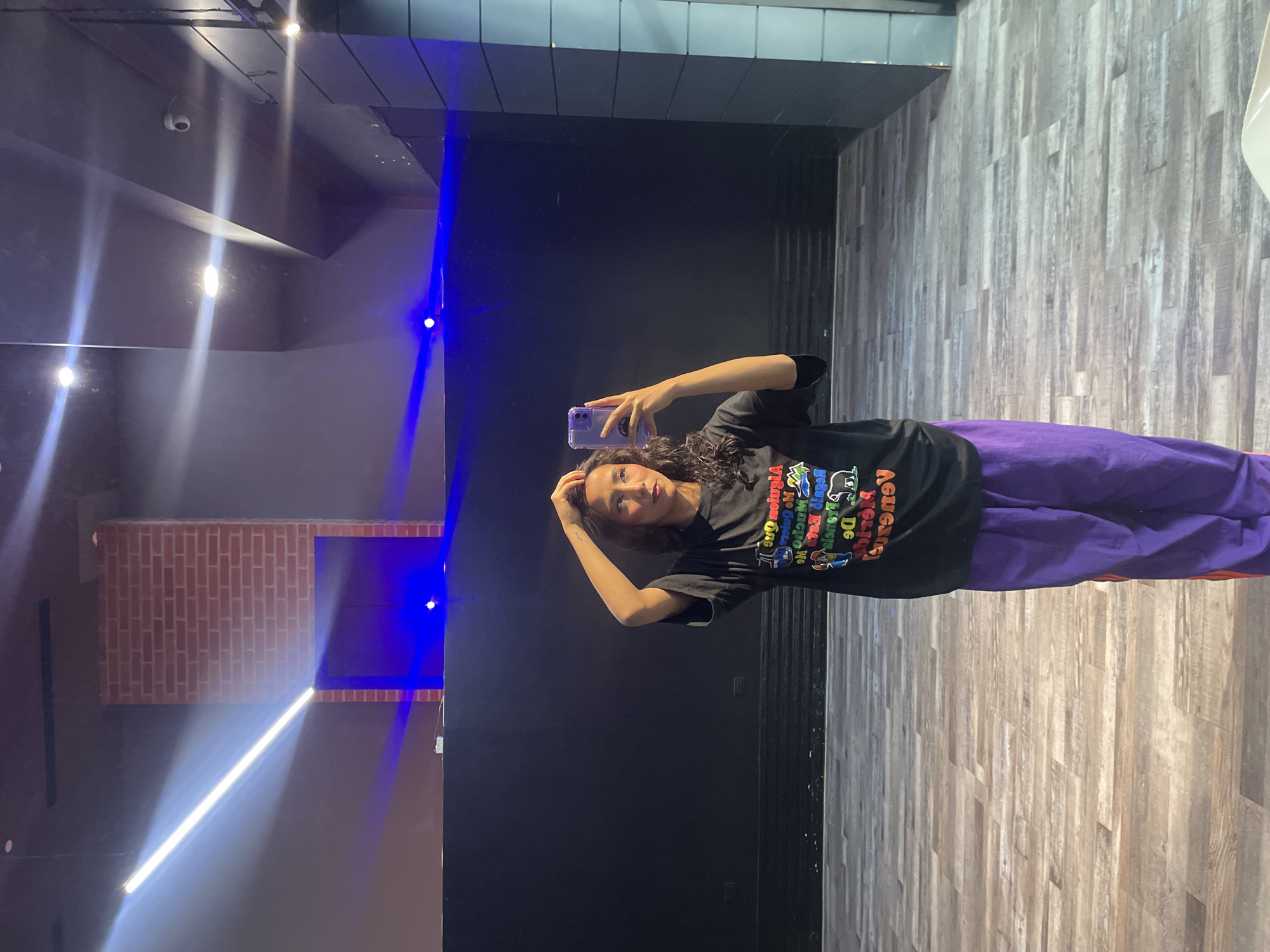 The image shows a young woman taking a selfie in a dance studio. She is wearing a black t-shirt with a colorful graphic print and purple pants. The studio has a dark backdrop, wooden floors, and dramatic lighting from blue and white LED strips. A brick-patterned wall is visible in the background. The woman is standing in front of a large mirror, holding her phone in her right hand and using her left hand to adjust her hair. The scene suggests a casual and energetic environment, possibly during a dance practice or rehearsal. The woman's pose and the setting imply an interest in dance or performance.