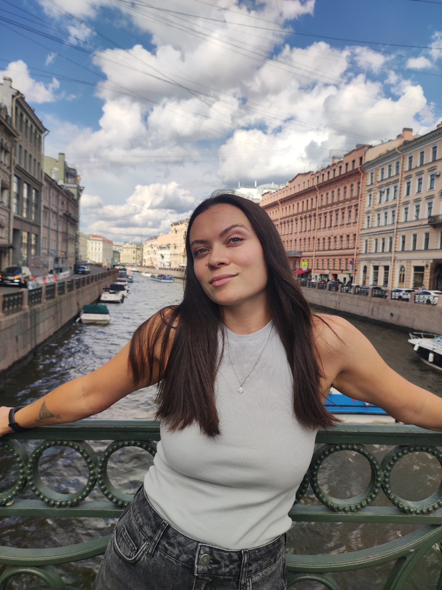A young woman with long dark hair and fair skin leans against a decorative green metal railing on a bridge overlooking a canal in Saint Petersburg, Russia. She is wearing a light grey ribbed tank top and dark grey distressed denim jeans. A small silver pendant necklace adorns her neck. Her right arm is bent and resting on the railing, with a black fitness tracker on her wrist. A tattoo is visible on her upper right arm. The canal is lined with buildings in various shades of orange and yellow, exhibiting classical architectural styles typical of Saint Petersburg. Several boats are docked along the canal, and a few cars are visible on the street behind the buildings. The sky above is blue with fluffy white clouds, and power lines are strung across the upper portion of the image.