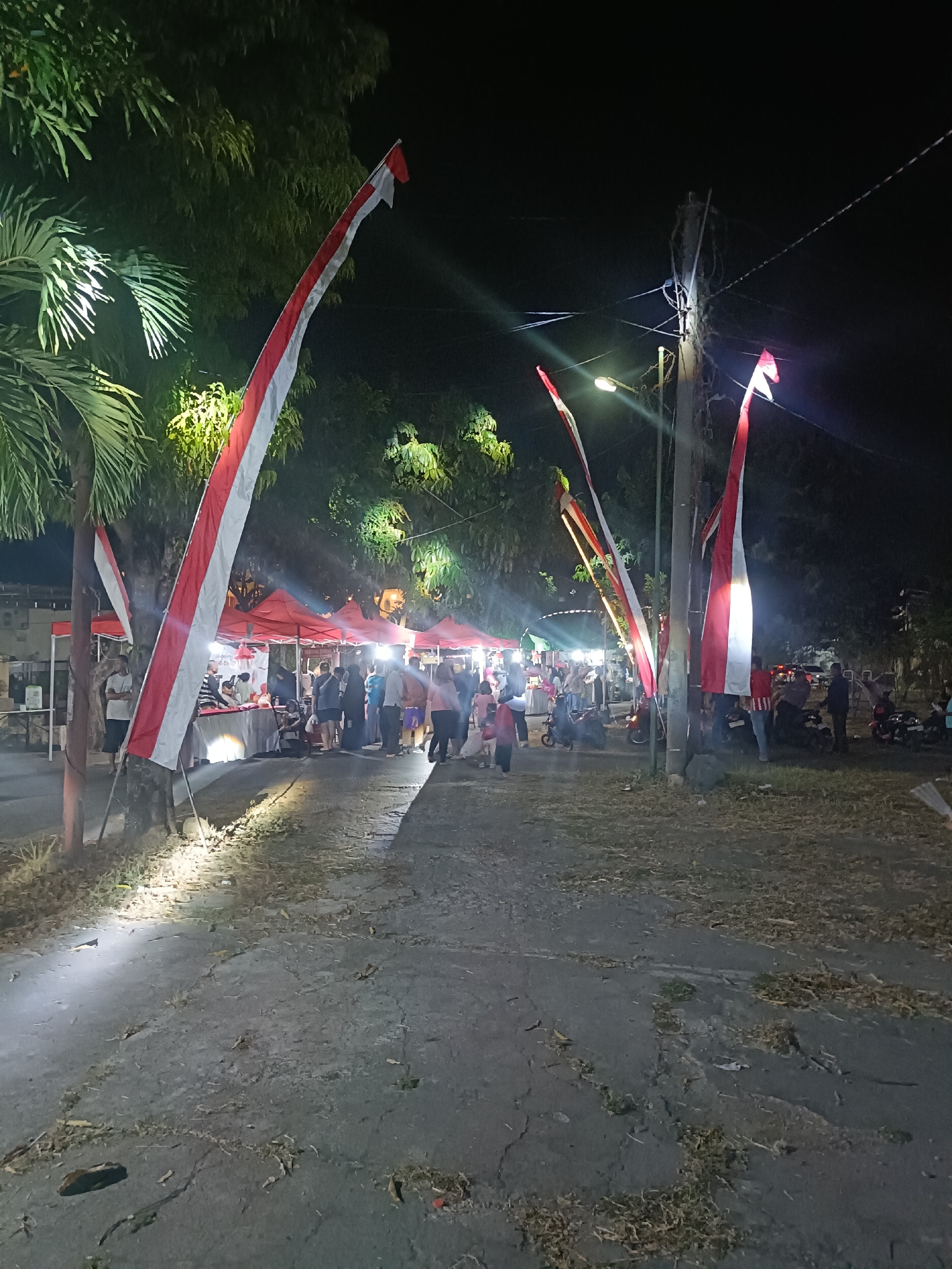 The image depicts an outdoor night market scene in Kediri City, Indonesia. A pathway, partially paved and cracked, leads into a gathering of people. Red and white Indonesian flags are prominently displayed, some attached to poles and others fluttering from what appear to be makeshift structures. Trees with lush green foliage border the pathway, and artificial lighting illuminates the scene, casting shadows and highlighting the path. Several red tents are set up, likely housing vendors. People are seen walking, standing, and interacting. Motorcycles are parked in the background. The atmosphere suggests a lively local event or festival.