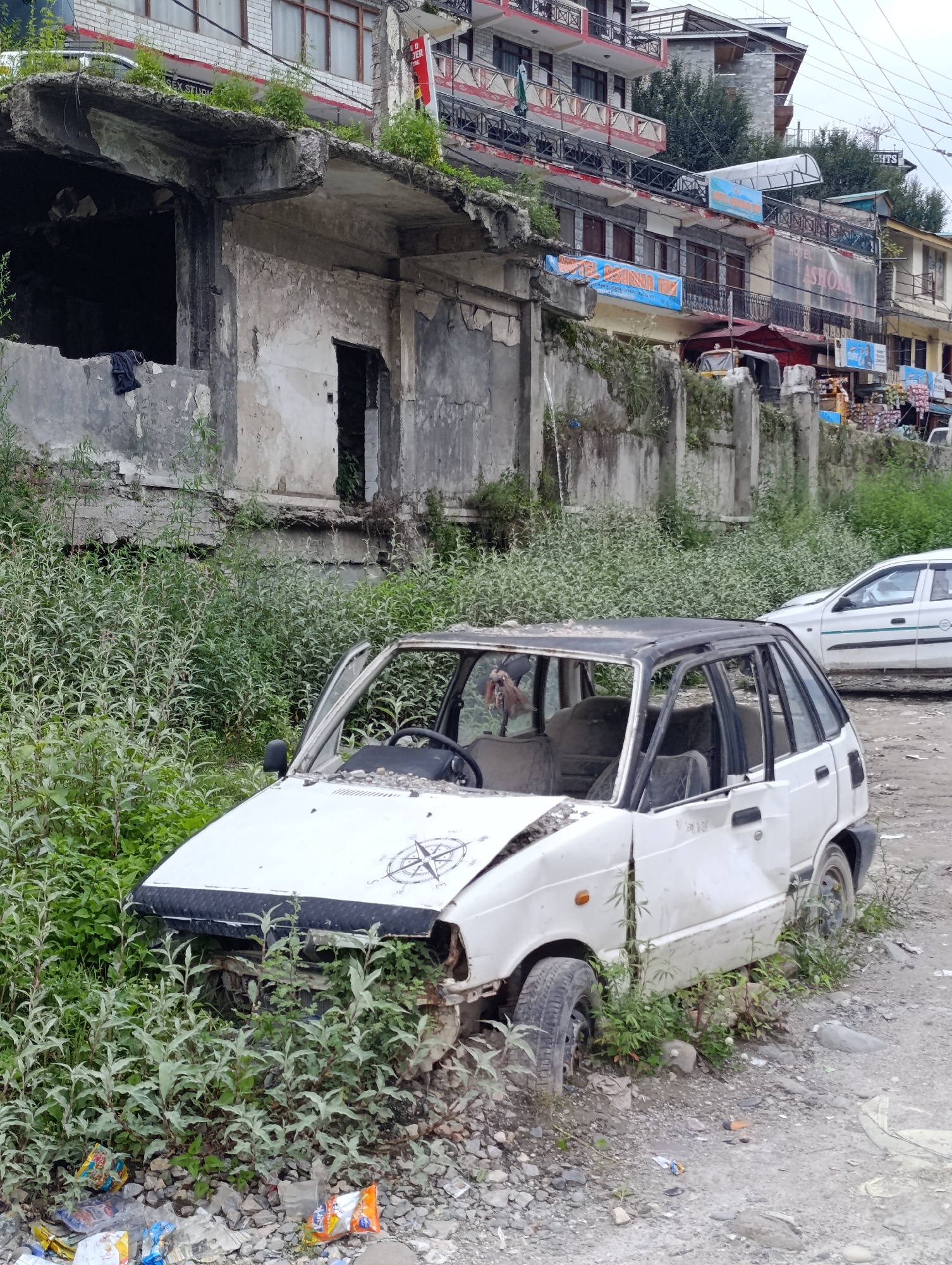 In Siumsa, India, a white, dilapidated Maruti 800 car sits abandoned amidst overgrown green vegetation and debris. The car's doors are open, and its windows are missing, revealing a damaged interior. The vehicle appears to be heavily weathered and has sustained significant damage, including a cracked windshield and rusted body panels. The surrounding area is a mix of dirt, gravel, and scattered trash, suggesting a neglected urban or semi-urban environment. In the background, multi-story buildings with balconies and advertising boards are visible, indicating a populated area. The overall scene portrays a state of disrepair and abandonment.