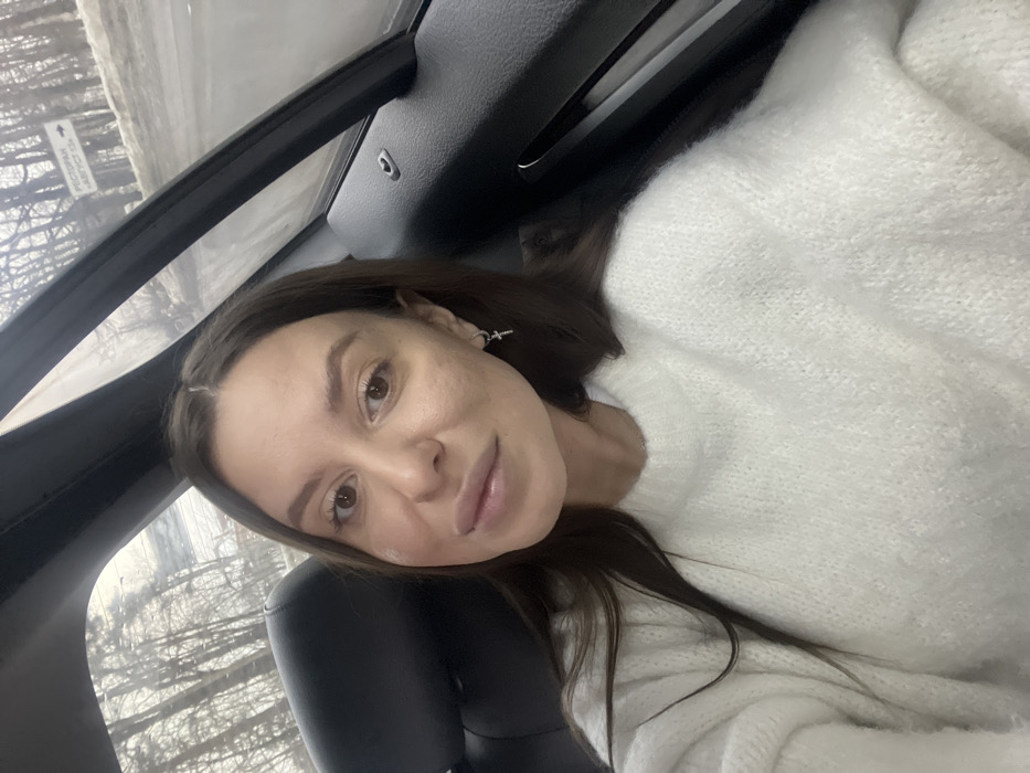 The image displays a female individual situated within a vehicle in Vysokovo, Russia. She is directly facing the camera, characterized by dark brown hair, light brown eyes, and full lips. She wears a white, knitted crew-neck sweater and a silver hoop earring with a cross pendant in her right ear. The vehicle's interior shows a black car seat headrest behind her left shoulder and a dark vehicle door panel to her right. Through the window, an exterior scene depicts a leafless, wintry or early spring landscape with bare trees and a wet, slushy road. A white rectangular sign, bearing black Cyrillic text, is visible and translates to "RESTAURANT 'AUGUST' -> 1.8 KM".