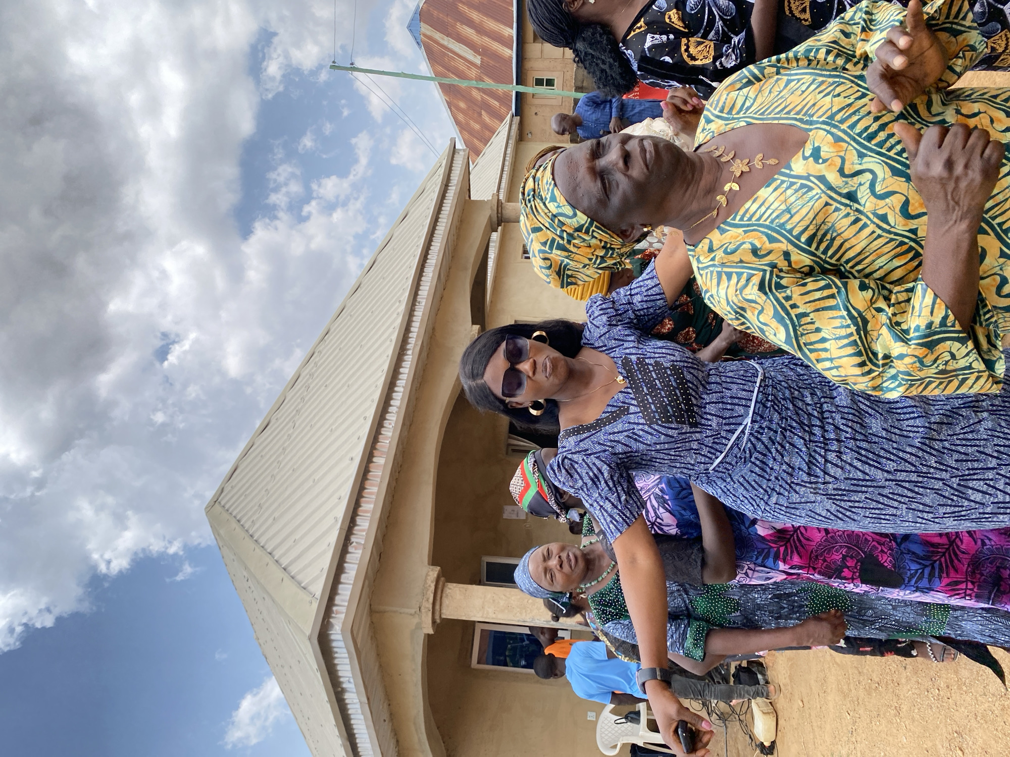 The image shows a group of people outdoors in Kuru Karama, Nigeria. A building with a light-colored, corrugated metal roof is visible in the background. The sky is partially cloudy with patches of blue. Several women are in the foreground wearing brightly patterned clothing. One woman is wearing sunglasses. There appear to be more people standing behind them, and a few people are seated on white plastic chairs. A few men are visible in the background wearing blue shirts.