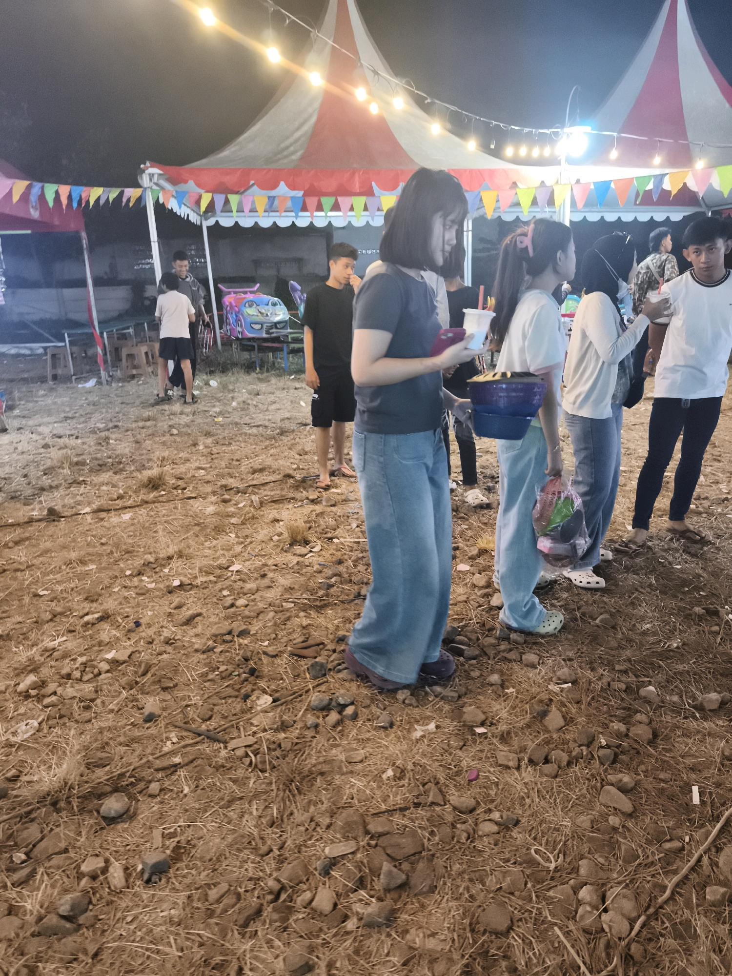 The image depicts an outdoor gathering at night, likely a fair or market, with a large tent in the background adorned with colorful pennant banners and string lights. The ground is covered in dry grass, small stones, and debris, suggesting an unpaved area.
In the foreground and mid-ground, several people are visible, mostly young adults and teenagers. On the left, a young woman with short dark hair, wearing a dark t-shirt and light blue wide-leg jeans, holds a phone in her left hand and a white cup with a red straw in her right. Next to her, another young woman with her hair tied back in a ponytail, wearing a light-colored t-shirt and light blue jeans, holds a stack of blue bowls. To her right, a person wearing a hijab and a white top is visible. Further to the right, a young man in a white t-shirt and dark pants holds a white cup. In the background, more individuals are present, including a young man in a black t-shirt and shorts.
Objects visible include the tent, string lights, pennant banners, plastic cups, bowls, and a plastic bag filled with items. There are also some tables and chairs partially visible in the background, and a small ride or attraction with a colorful cartoon character. The overall scene suggests a casual, festive atmosphere at an outdoor event.
The location is identified as Palembang, Indonesia. While the specific area within Palembang is not detailed, the setting suggests a community event or market.