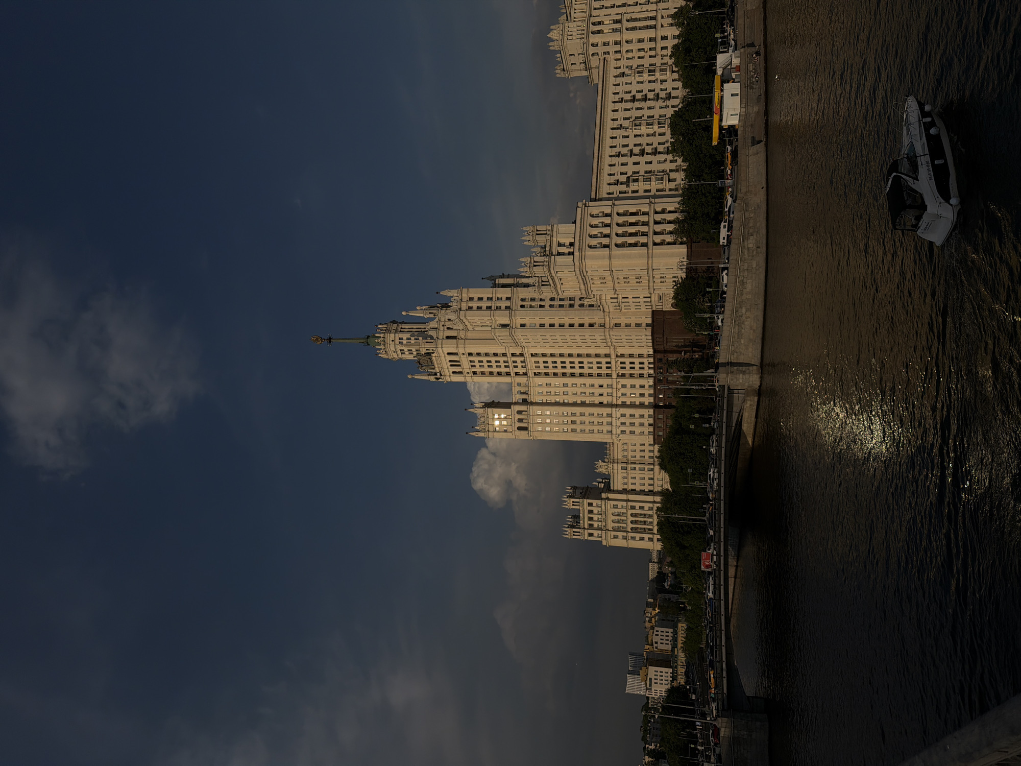 The image shows a view of Kotelnicheskaya Embankment Building in Moscow, Russia, along the Moskva River. The skyscraper is prominently featured against a dark blue sky with some clouds. Along the river embankment, there are trees and parked cars. A boat is visible on the water.