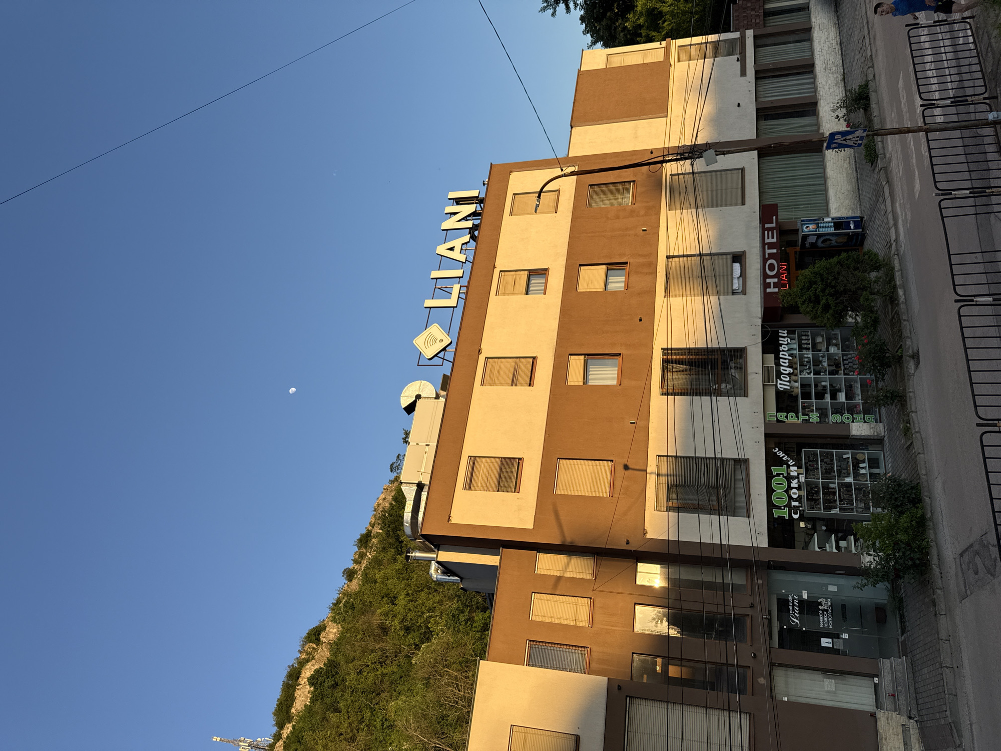 The image shows a building in Lovech, Bulgaria, identified as the "Liani" Hotel by the prominent sign on its roof. The building has a brown and beige facade with multiple windows. Below the hotel sign, a circular logo is visible. On the ground level, there are shops, including one called "1001 Стоки" and another with a sign in Cyrillic possibly translating to "Gifts". Another sign says "Piano Hair Studio". A street is visible to the right of the building with metal safety barriers alongside. A small pedestrian crossing sign can also be seen. Behind the building on the left is a hill covered in dense vegetation. A clear blue sky with a faint moon is visible above. A power line stretches across the upper portion of the sky. The photo is rotated counter-clockwise.