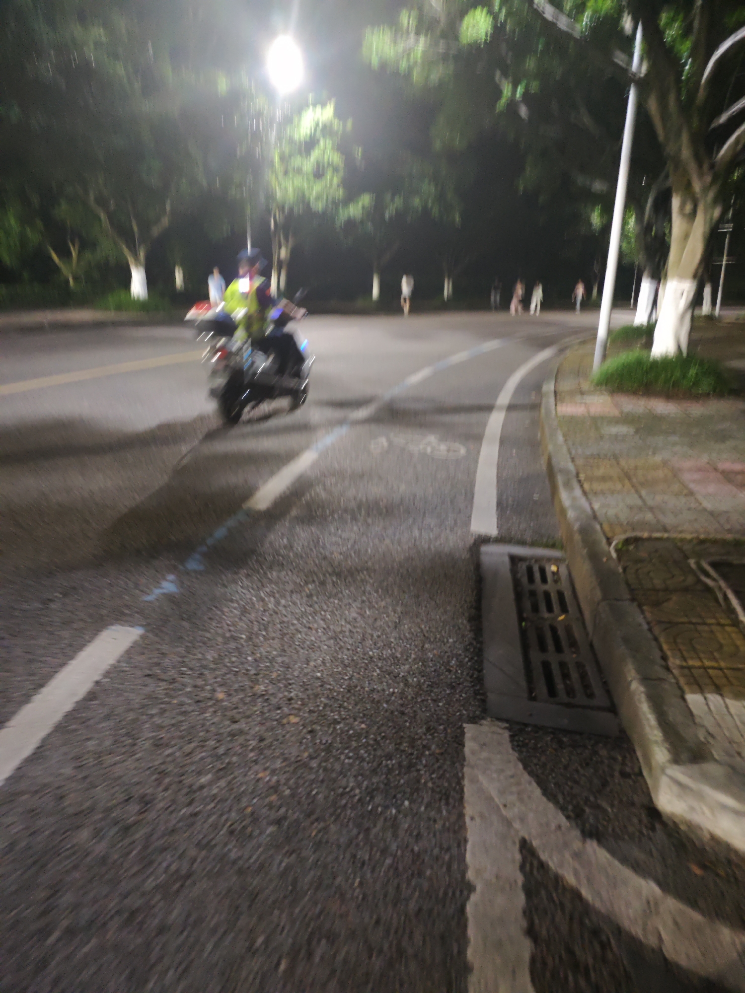 A blurry, nighttime scene shows a person on a motorcycle moving from left to right on a wet asphalt road. The person is wearing a bright yellow vest and dark clothing. The road has a white line marking its edge and a blue marking further into the lane. To the right of the road is a concrete sidewalk with a drainage grate. Trees with white-painted bases line the area beyond the sidewalk. In the background, several indistinct figures are visible walking. The scene is lit by a bright overhead streetlamp. The location is the Chongqing High-tech Zone, China.