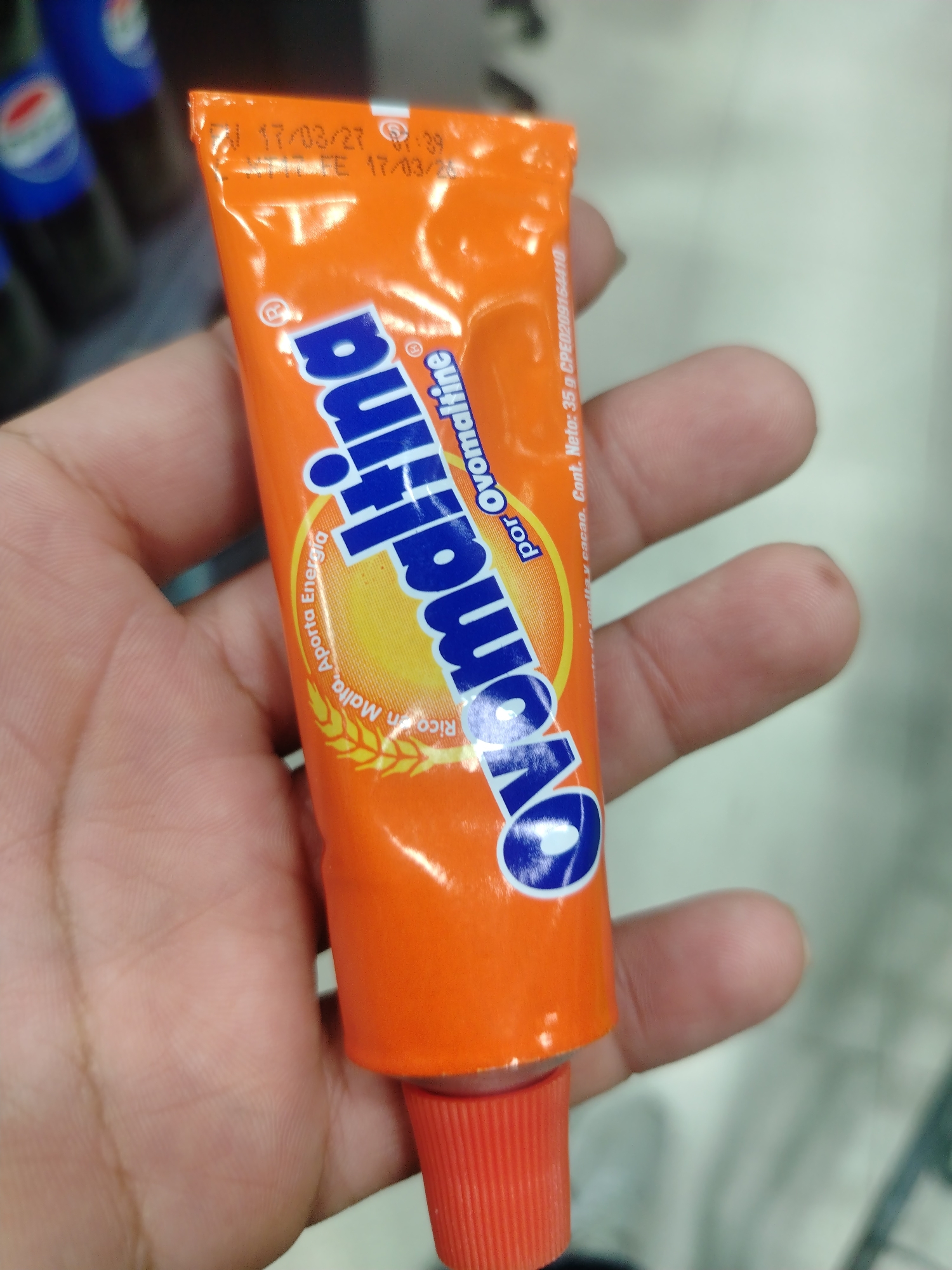 An orange tube of Ovomaltine is held in a person's left hand, with fingers partially obscuring the tube. The tube has blue lettering that spells out "Ovomaltine." Text on the tube indicates production or expiration dates, with "17/03/27" and "17/03/25" visible. There is also text stating "Cont. Neto: 35 g CPED209154410." The background is blurred, showing what appears to be shelves with blue containers, suggesting a retail environment, potentially a supermarket or pharmacy in Juan Griego, Venezuela.
