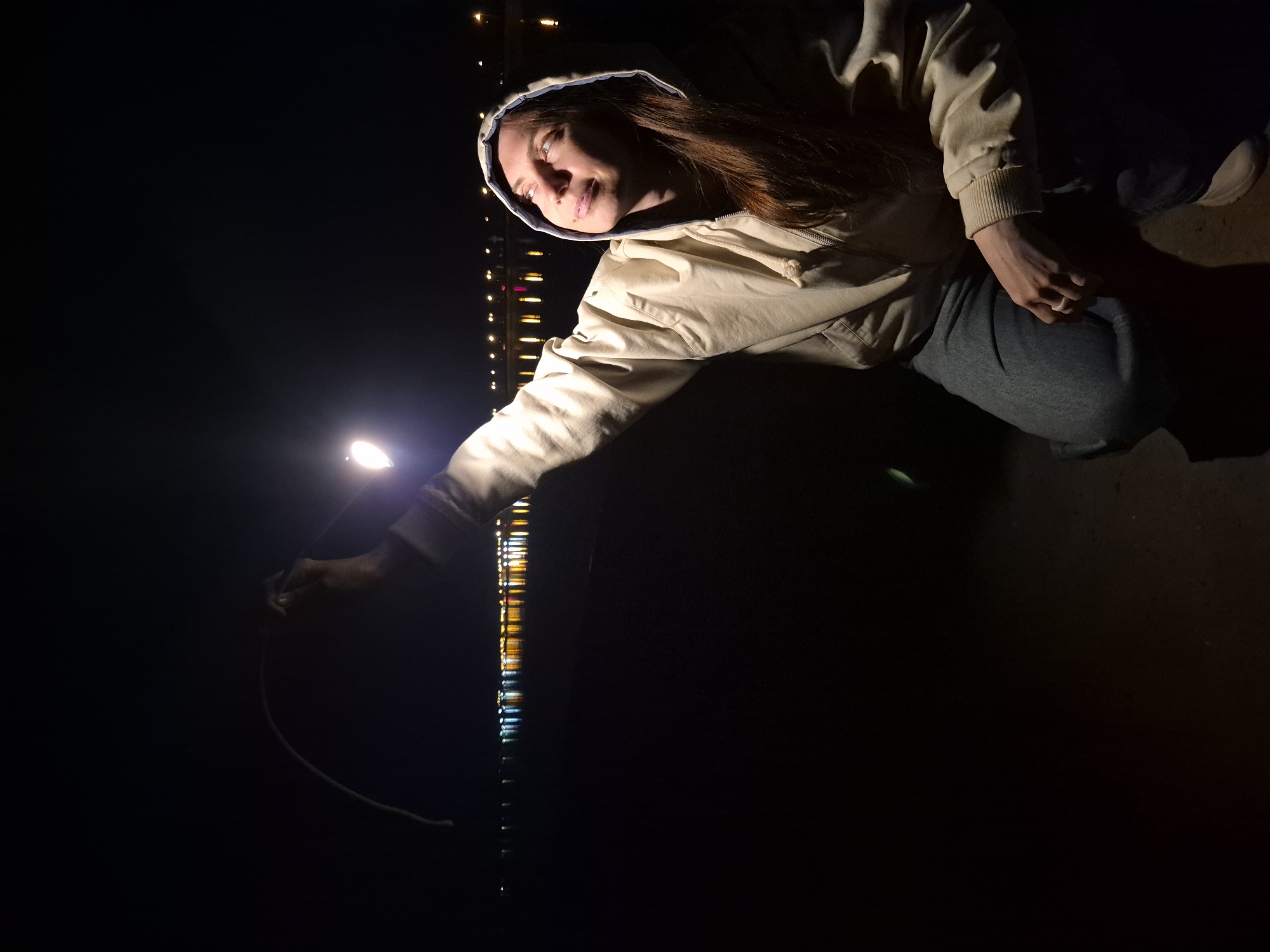 At night in Unknown, Unknown, a person wearing a hooded jacket and jeans is kneeling on a dark surface. The person extends their arm holding a lit object. In the background, a line of lights is visible along the edge of what appears to be a body of water. The scene is dark, with the primary source of light emanating from the object held by the person.