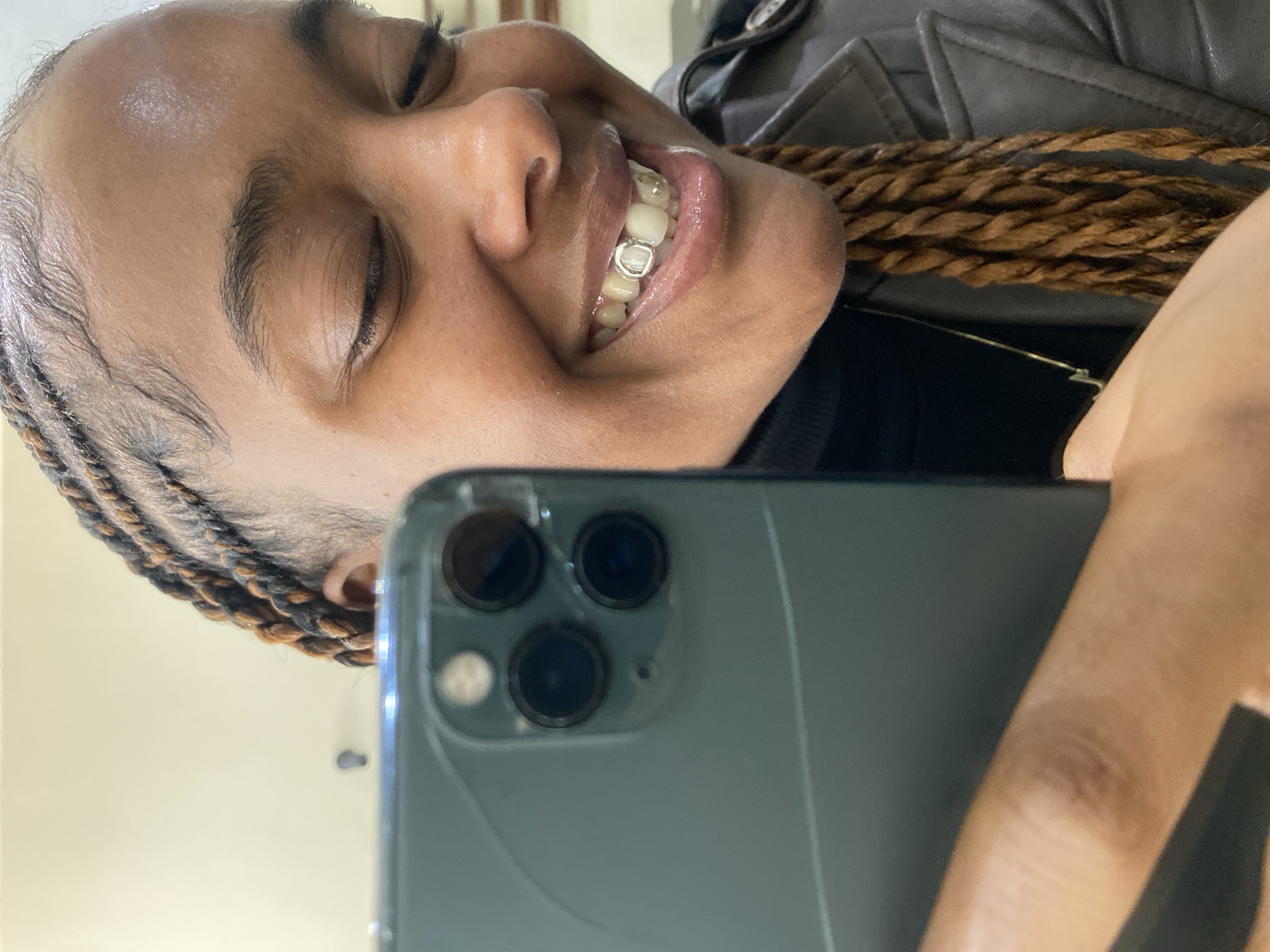 An angled mirror selfie captures a person, likely a woman, with their head tilted, smiling broadly with closed or squinting eyes. She has a dark skin tone, with braided hair featuring light brown or blonde highlights and meticulously styled baby hairs along her forehead.
Her wide smile reveals white teeth, with metallic caps (grillz) visible on at least two upper teeth, specifically one canine and one incisor. Her lips appear glossy. She is wearing a dark top, possibly a turtleneck, and a dark jacket that appears to be made of leather. A thin gold-colored chain or necklace is visible around her neck.
The person is holding a dark grey or green smartphone, which is prominently featured in the lower right and center of the frame. The phone's back is visible, showing a triple camera system and a flash, indicative of a recent iPhone model. The background behind the person is indistinct and light-colored, suggesting an indoor setting. The overall expression conveys happiness or contentment. No text is visible in the image. The location context provided is London, United Kingdom.