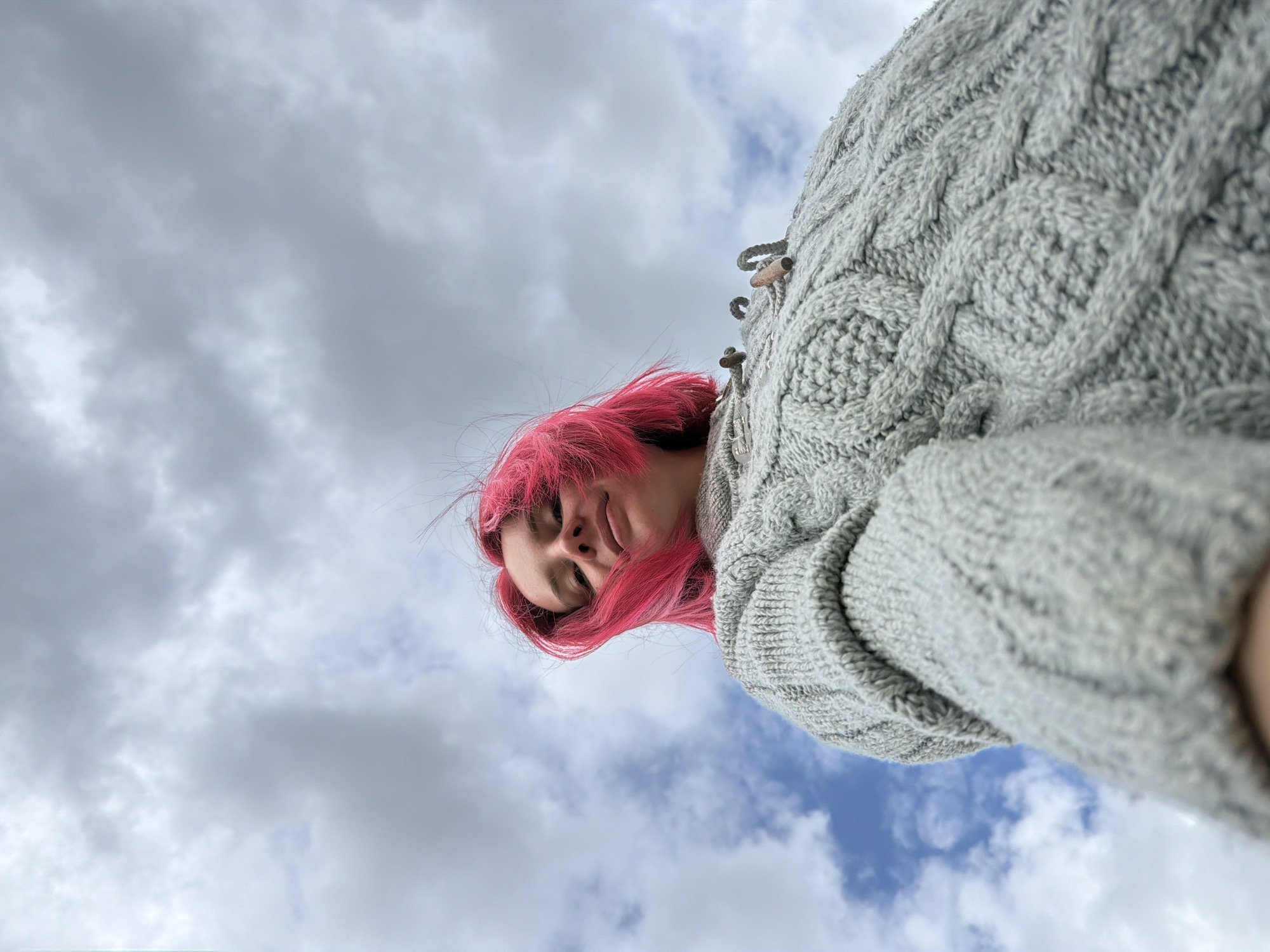 A low-angle shot depicts a person with vibrant pink hair wearing a grey cable-knit sweater. The person is looking down at the camera with closed eyes. Behind the person is a sky filled with grey and white clouds. The specific location in Chaikovsky, Russia, cannot be determined from the image alone.