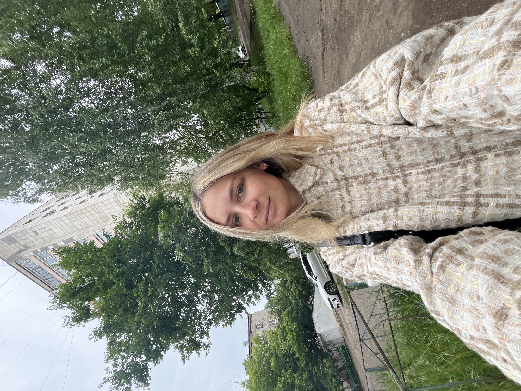 A selfie captures a blonde woman outdoors in what appears to be a residential area of Chaikovsky, Russia. The woman, likely in her 20s or 30s, is positioned in the foreground, looking directly at the camera with a neutral or slightly smiling expression. Her long, blonde hair frames her face, and she has fair skin and blue eyes. She is wearing a textured, tweed-like jacket or coat with a woven pattern in shades of cream, beige, and light brown. The garment has visible buttons on the front, and a dark strap, possibly from a bag, crosses her torso.
The background reveals a green and urban landscape. Dominating the upper and middle sections are lush green trees with dense foliage, suggesting late spring or summer. Patches of green grass are also visible. To the left, a tall, light-colored apartment building with multiple windows, some featuring pink sills, rises into the frame. Further back, another, smaller grey building with white windows can be seen. A white car is partially parked on a paved surface, which appears to be a road or path. The sky overhead is overcast and light grey. There is no visible text in the image. The scene conveys a casual, daytime moment in a typical urban environment.