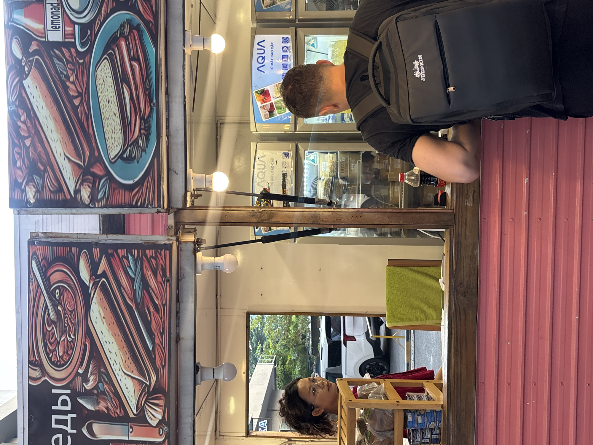 The image shows a street food stall with two illuminated signs depicting food items. In the foreground, a man wearing a black t-shirt and backpack leans over the counter. Behind him, a young woman with glasses and curly hair is looking out from behind the counter. The stall appears to be open, with various food items and refrigerators visible inside. The exterior of the stall is painted pink. In the background, a white SUV is parked on the street. The scene is illuminated by natural light and the stall's lighting.