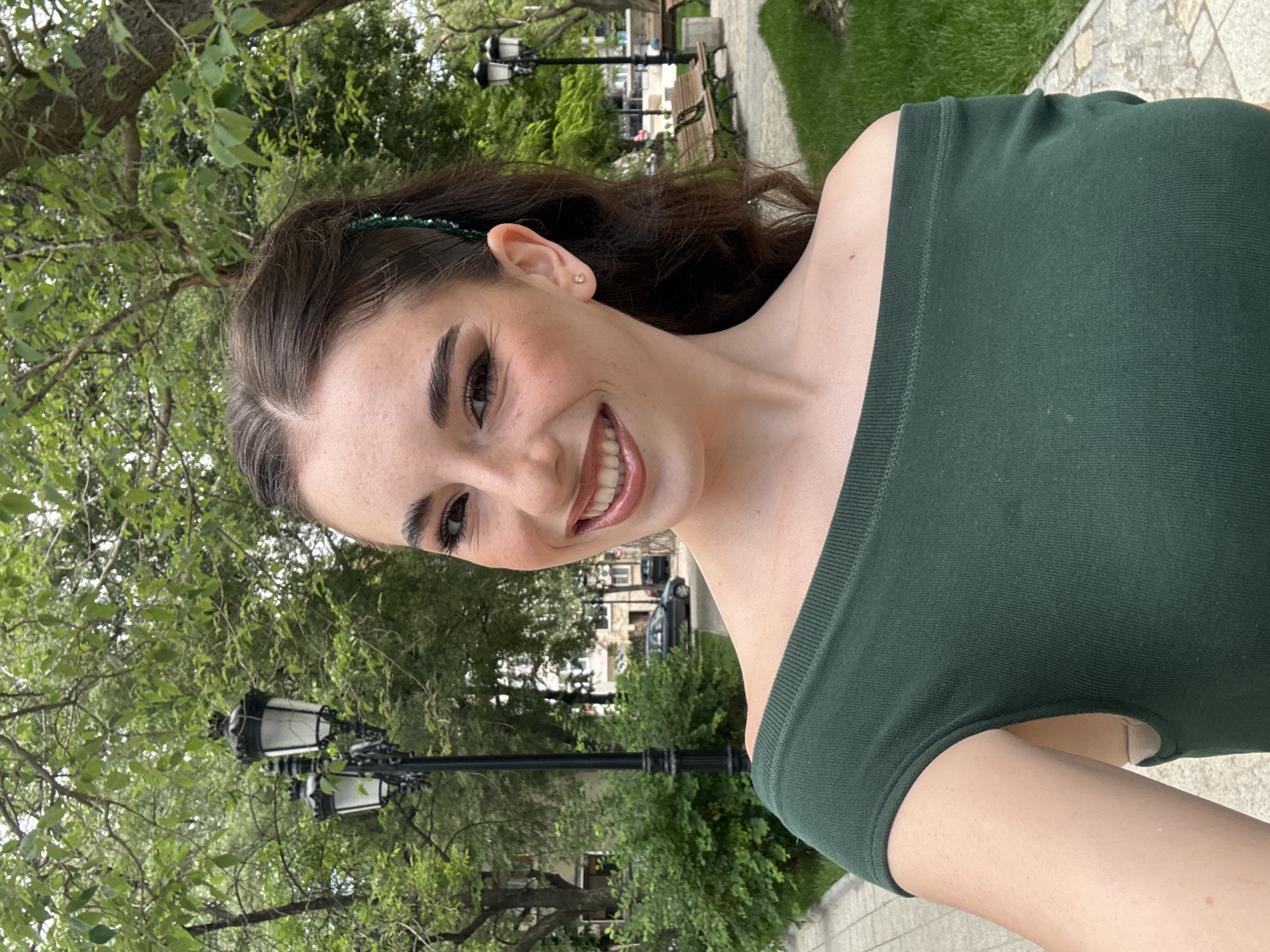 The image is a selfie taken outdoors in a park or garden-like setting. The subject is a young woman with dark hair and fair skin, wearing a dark green off-the-shoulder top. She is smiling and looking directly at the camera. Her hair is pulled back, and she has a green beaded headband. The background includes lush greenery, trees, lampposts, benches, and a stone-paved path. Buildings are visible in the distance, suggesting an urban environment. While the image doesn't explicitly confirm it, the lampposts, greenery, and architecture are consistent with features found in the city of Plovdiv, Bulgaria. However, a definitive location within Plovdiv cannot be determined from the image alone.
