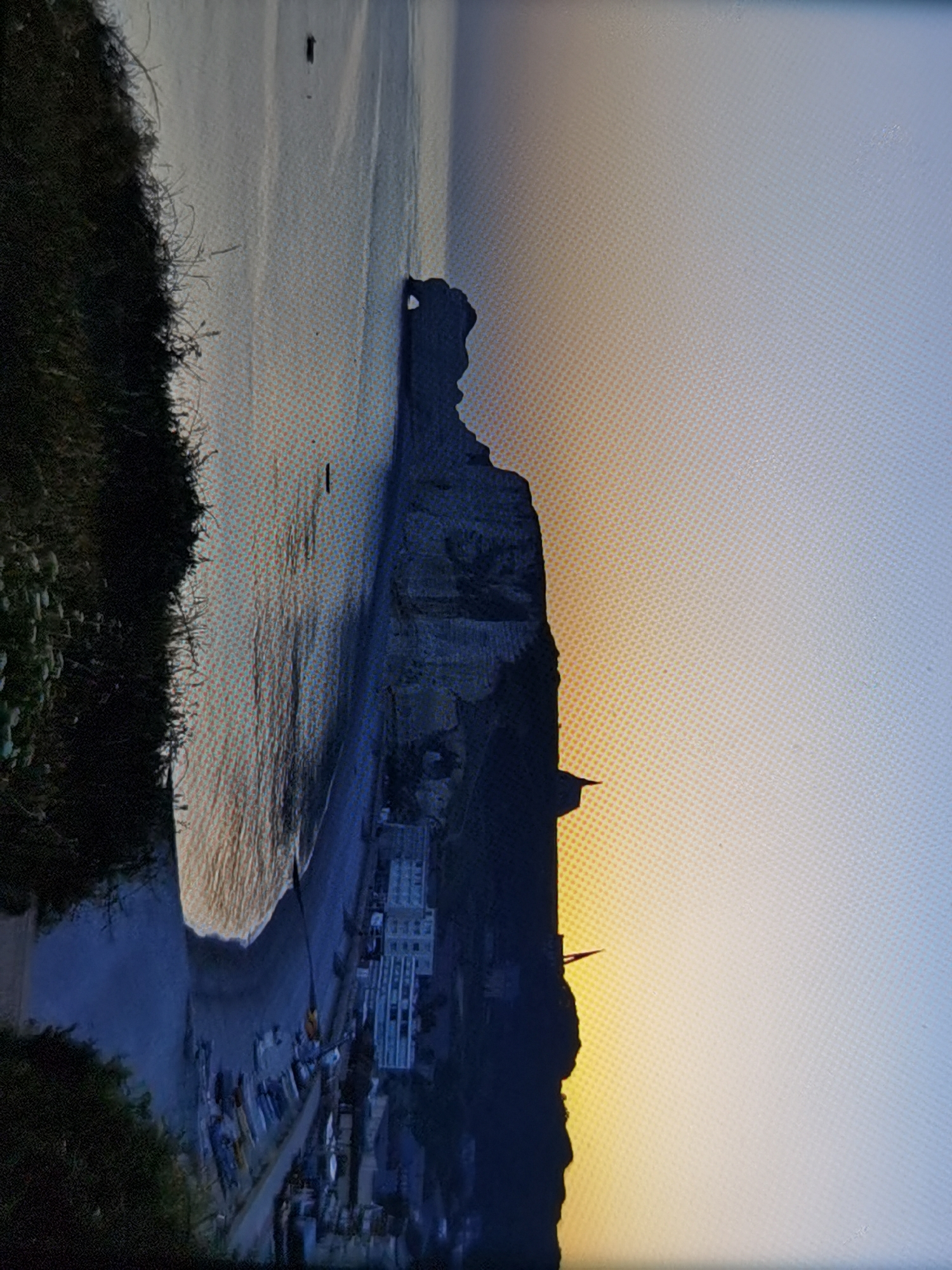This media file depicts a scenic view of the cliffs of Étretat, France, during sunset. The coastline features a prominent rock formation that juts out into the sea, silhouetted against the warm hues of the setting sun. Below the cliffs, a small town is nestled along the shore, with buildings and boats visible. The beach appears to be a mix of sand and pebbles.
The time of day is clearly sunset, as indicated by the gradient of orange and yellow in the sky, transitioning to a pale blue. The weather appears calm, with still water reflecting the sky. The overall mood is serene and picturesque. There is no visible text in the image.