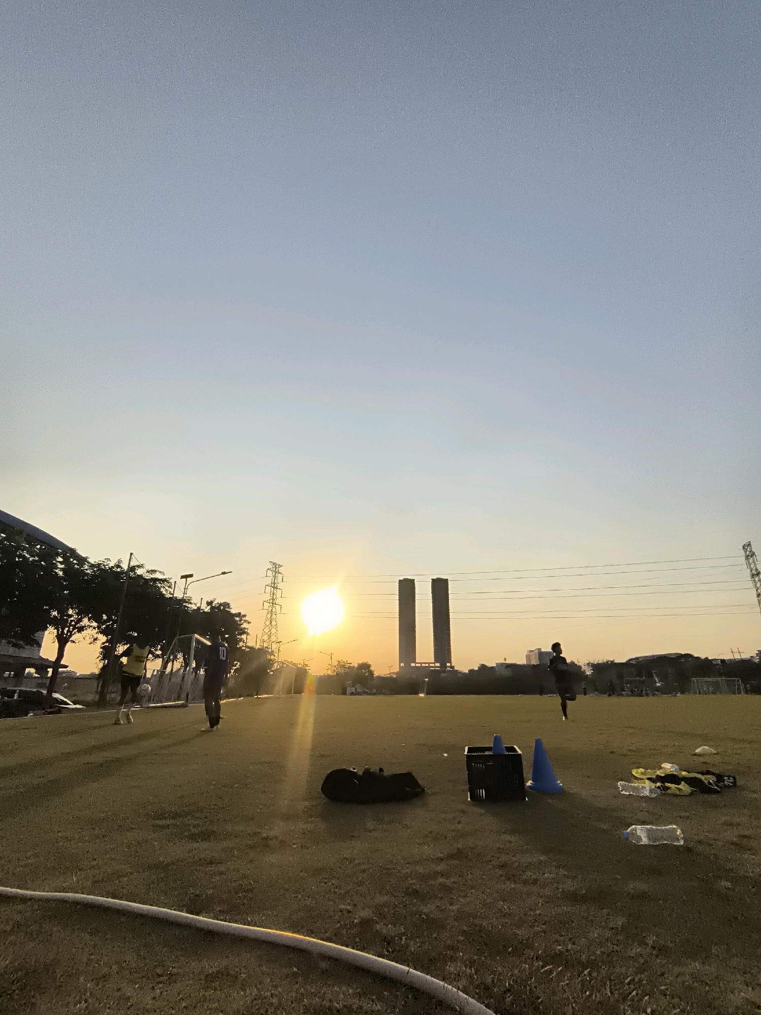 An outdoor, wide-angle, low-angle shot captures a soccer field at sunset, with the sun low on the horizon. The sky is a gradient of light blue and soft orange. In the distance, two identical, dark, rectangular buildings stand tall. A high-voltage electrical tower with crisscrossing wires is visible to the right of the buildings. Two individuals are on the field. On the left, a person in a yellow shirt appears to be interacting with a soccer ball, while another person in a dark shirt stands nearby. Further in the distance, a lone figure in dark clothing runs across the field. Scattered on the grass are various objects, including a black bag, a blue plastic cone, a black crate, discarded water bottles, and yellow and black sports apparel. The foreground shows a large, white hose coiled on the grass. The image appears to be taken in Surabaya, Indonesia, evidenced by the presence of similar architectural structures and the general atmosphere of a sports facility.