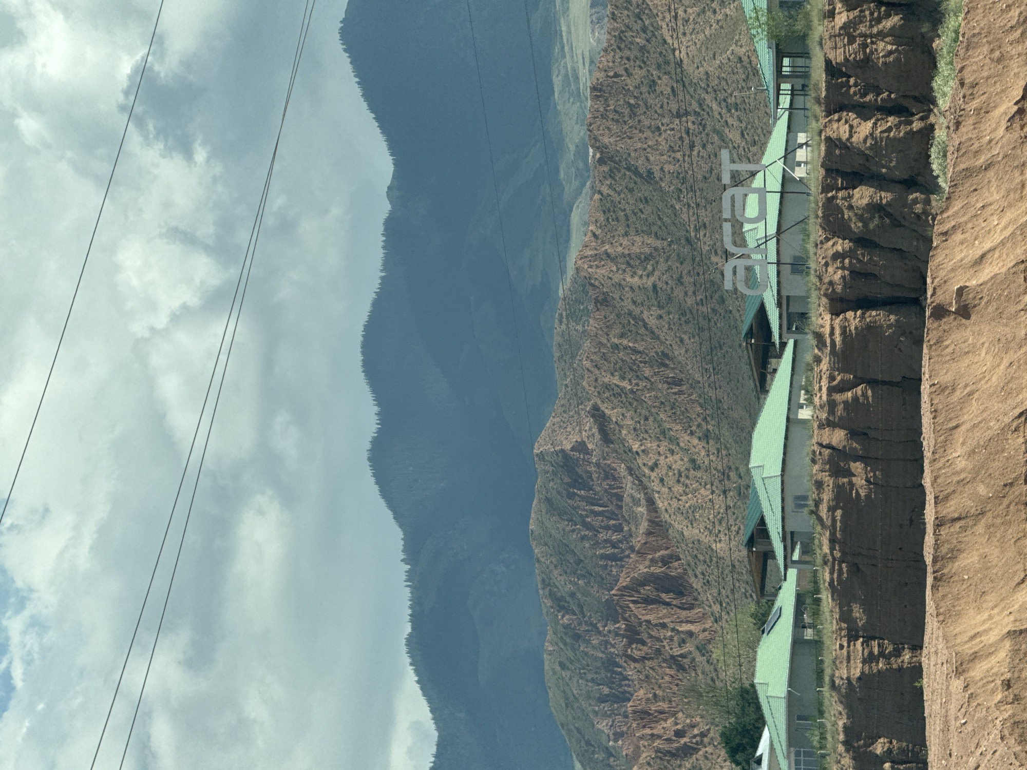The image depicts a scenic view of Kaji-Say, Kyrgyzstan, with a prominent natural landscape and man-made structures. In the foreground, a reddish-brown cliff face, characteristic of the region's geology, dominates the lower portion of the frame. Above this, a cluster of modern houses with distinctive bright green roofs are visible, situated on a plateau. These houses appear to be part of a settlement or resort, with some exhibiting solar panels on their rooftops. In the background, a series of layered mountains rise majestically. The middle mountains are rugged and reddish-brown, with sparse vegetation, while the furthest mountains are larger, covered in dark green coniferous forests, and shrouded in a misty, blueish haze. The sky is partly cloudy, with large, soft white clouds against a pale blue backdrop. Overhead, power lines are strung across the scene, suggesting the presence of infrastructure. A large, stylized white sign with the letters "ARAT" is prominently displayed above the houses, likely representing a business or a place name. The overall impression is one of a tranquil, natural setting complemented by modern development.