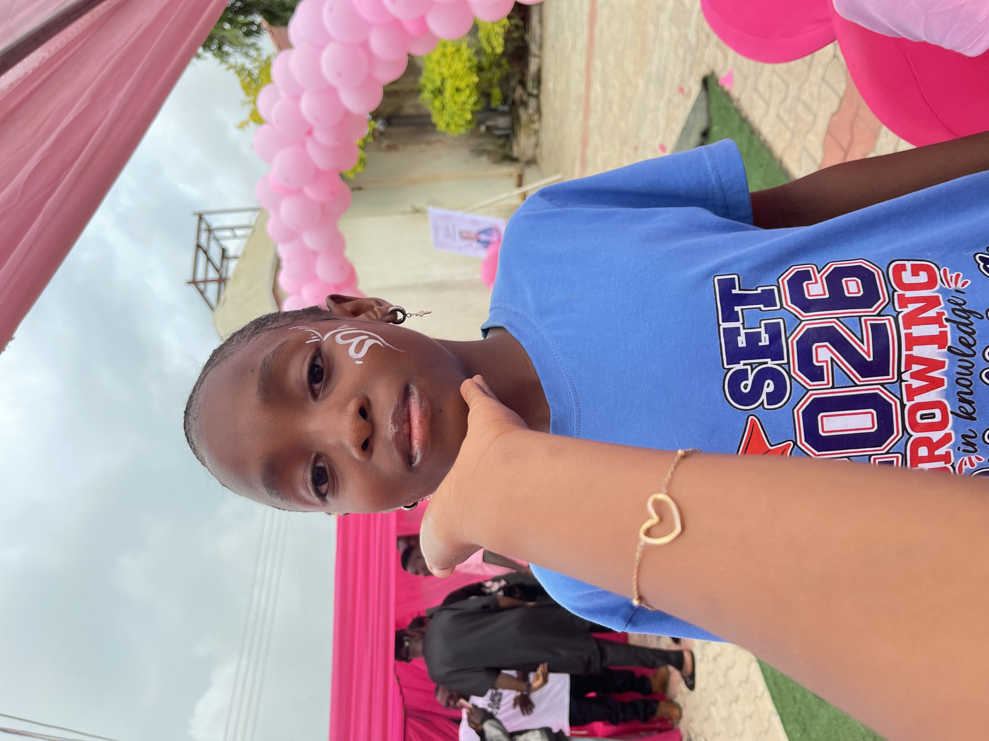 A medium shot captures a young Black person from the chest up, facing the camera in an outdoor setting. The subject has dark skin and hair, and is wearing a blue t-shirt with white, red, and blue lettering that reads "SET 26 GROWING in knowledge". White face paint is applied to the left side of their face in swirling patterns, and they are wearing a black hoop earring with a small charm on their left ear. A hand with a gold bracelet with a heart charm is held up to the subject's chin. In the background, pink balloons are arranged in an arch, and pink fabric drapes from a structure overhead. Other people are visible in the background, including one person wearing a black traditional outfit and another in a white t-shirt. The location appears to be Gura Top, Nigeria, given the context. The ground is covered with green artificial turf and paved tiles. The sky is overcast.