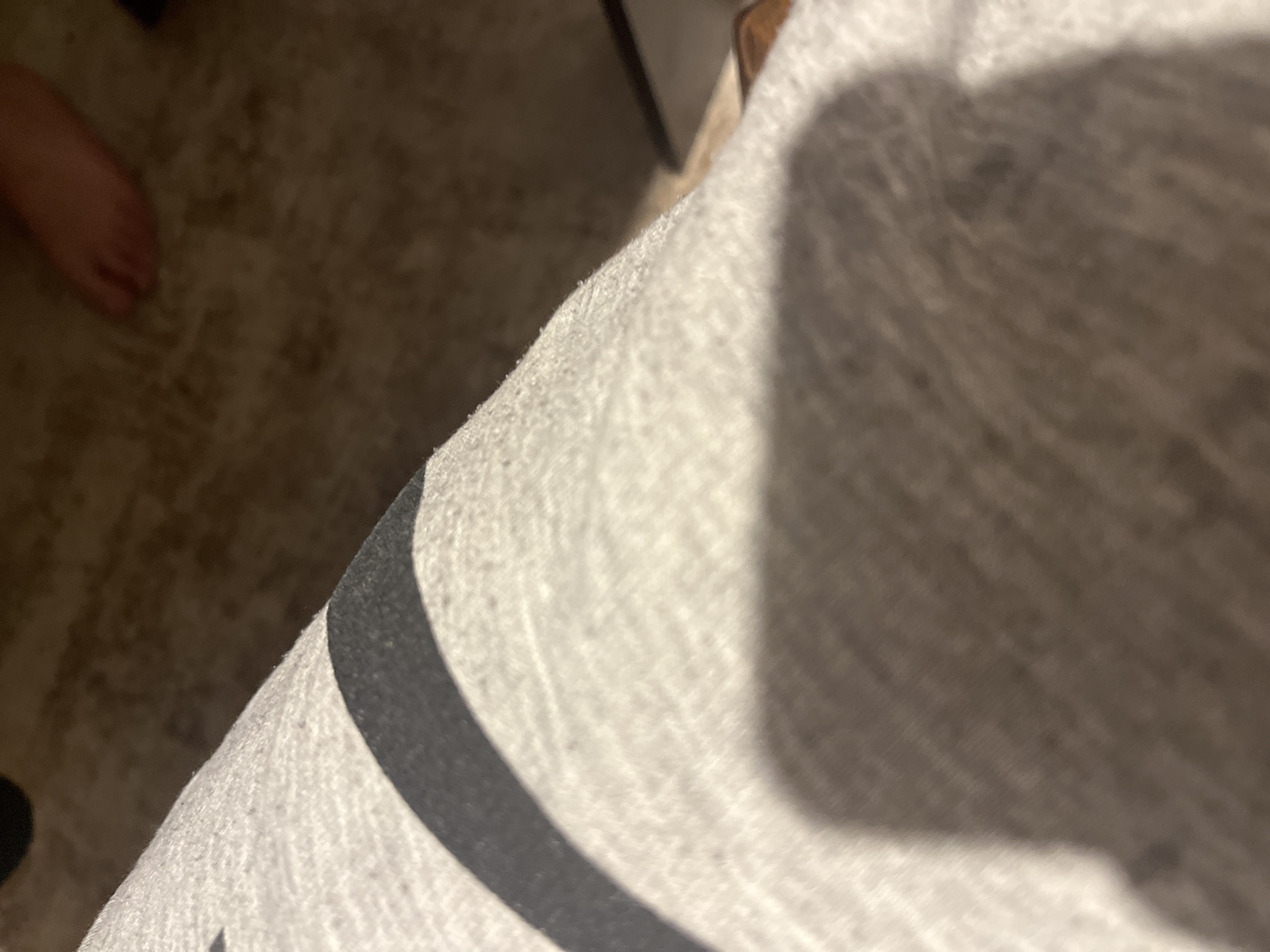The scene is located in Blagoevgrad, Bulgaria. The image presents an angled, close-up view of light-colored, textured fabric, likely a garment, featuring a dark, curved stripe. A distinct shadow obscures a significant area of the fabric. In the blurred background, a brown, textured surface, consistent with carpeting, is visible. A partially discernible, bare human foot is present in the upper right quadrant of the background, indicating the presence of an individual within an indoor setting. No active interaction or specific action is depicted.