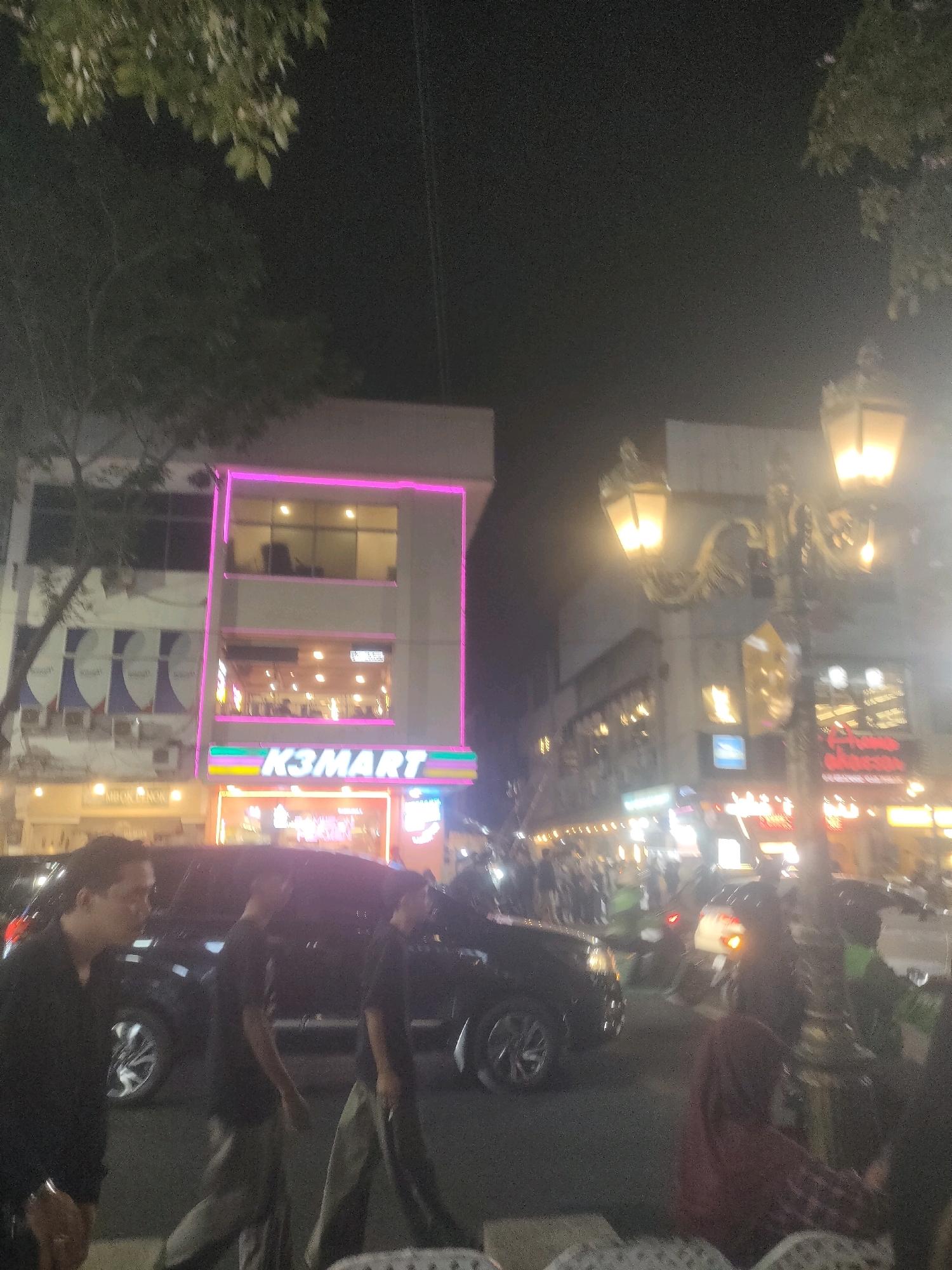 The image captures a bustling street scene at night, likely in Surabaya, Indonesia, as indicated by the "K3MART" sign, a common convenience store chain in Indonesia. The foreground is dominated by the silhouettes of several individuals walking and standing near parked vehicles. A dark SUV is prominent, with its headlights and taillights possibly visible.
The environment is illuminated by the warm glow of streetlights and the bright neon lights of businesses lining the street. A two-story building with pink neon accents is clearly visible, with "K3MART" displayed on its facade. The upper floor has windows reflecting the interior lighting, while the lower floor appears to be a storefront. Further down the street, a decorative lamppost with multiple lamps adds to the ambiance. Other businesses with illuminated signage are visible in the background, and a crowd of people can be seen further down the street, suggesting a lively commercial or entertainment area. The presence of trees with visible leaves overhead indicates an outdoor setting. The overall impression is one of a vibrant, urban night life.