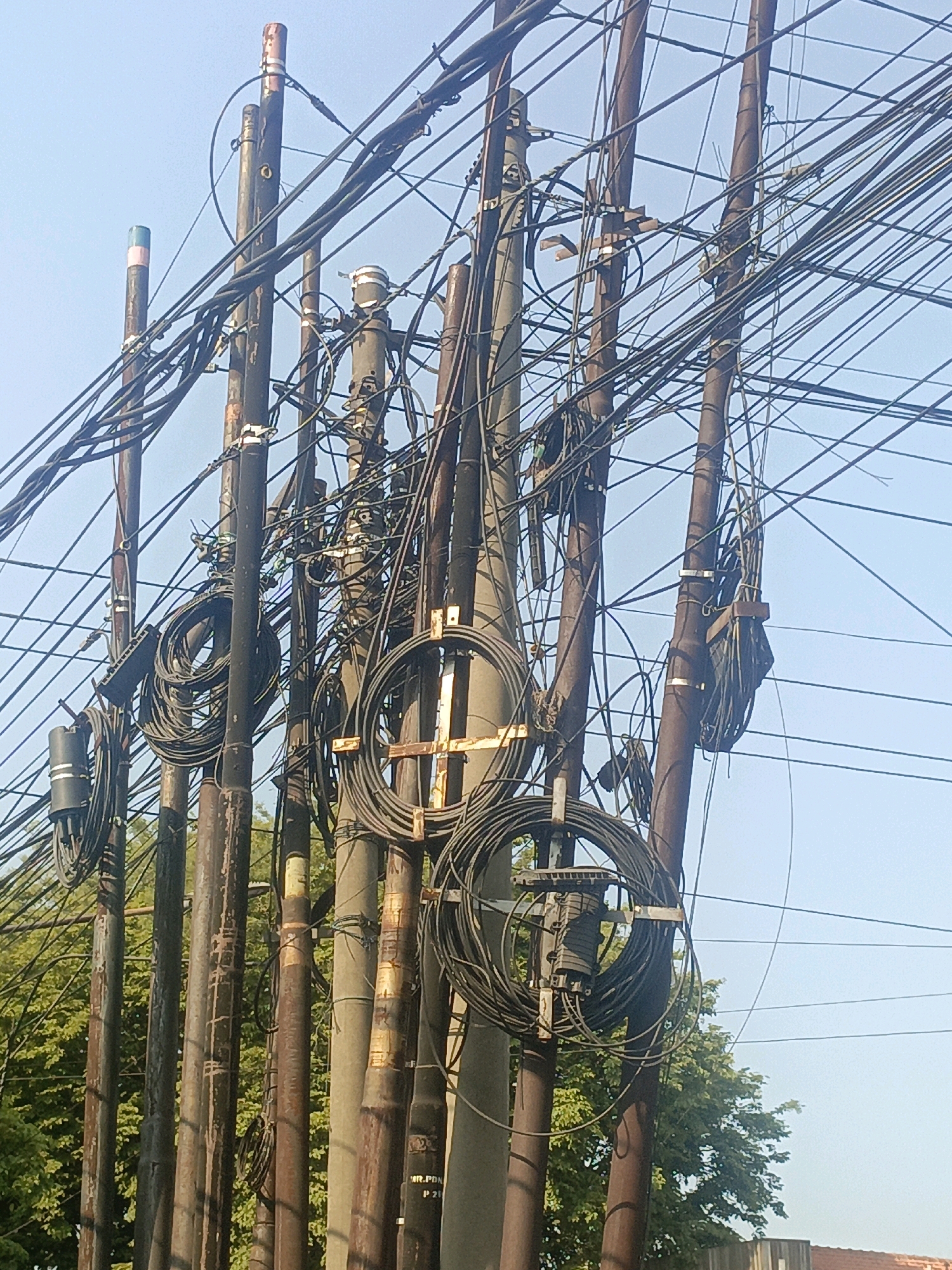 The image shows a cluster of utility poles entangled with a dense network of wires and cables against a clear blue sky. Several poles are visible, made of various materials including metal and concrete, with some showing signs of rust and wear. Wires are coiled around some poles and suspended between them, creating a chaotic and complex web. There are also some circular bundles of cable secured to the poles. In the background, green foliage and trees are visible, suggesting an outdoor, possibly semi-urban or rural, environment. A portion of a red tiled roof can be seen at the bottom right. The scene is static, with no people or discernible activity. The context provided specifies the location as Gempol, Indonesia.
