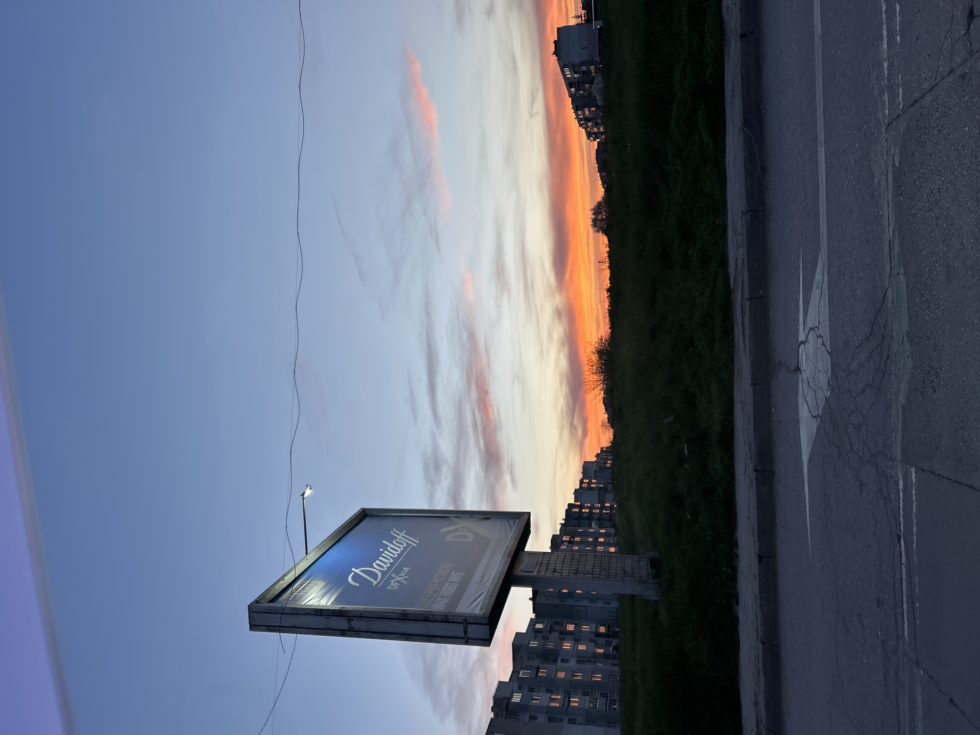 The image captures a street scene in Varna, Bulgaria, during a vibrant sunset. The sky transitions from blue to orange and pink hues. A billboard for Davidoff cigarettes stands prominently, illuminated by a street light. Behind it, a cluster of apartment buildings with lit windows is visible, suggesting early evening. A strip of green vegetation separates the buildings from the road. The road is dark gray asphalt with visible cracks and road markings. There are no people visible in the image.