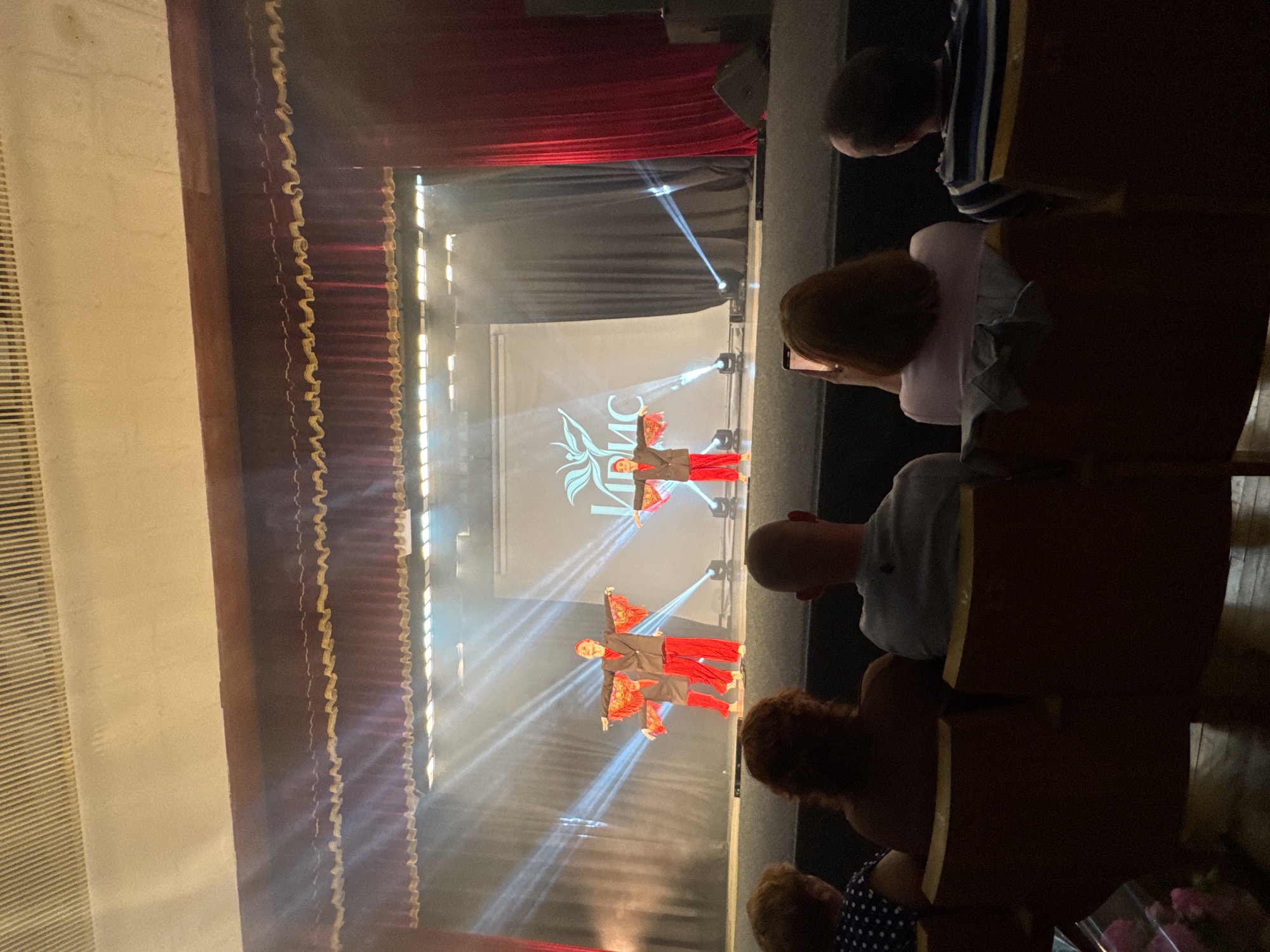 The image captures a performance on a stage, likely in Sochi, Russia, given the text on the screen. Four young dancers, dressed in red pants and black jackets with red decorative sleeves, are performing in front of a large screen displaying Cyrillic text that appears to read "ЛИРИС" along with a stylized logo of a bird. The stage is lit with spotlights, creating beams of light that cut through the smoky atmosphere. Dark red curtains frame the stage. In the foreground, the backs of an audience are visible, seated in dark chairs. One audience member in the center is holding up a smartphone, presumably to record the performance. The overall atmosphere suggests a theatrical event or dance recital.
