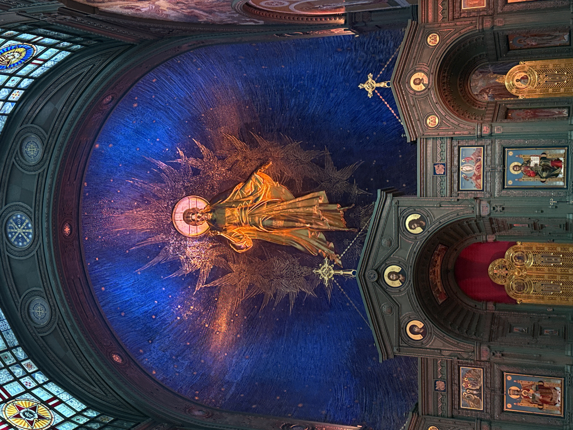 The image depicts the interior of a grand religious building, likely a church or cathedral. Dominating the scene is a large, golden statue of Jesus Christ, positioned centrally within a vibrant blue, star-speckled mosaic arch. The statue is illuminated, and rays of light emanate outwards from behind it. Below the statue, a golden, ornate cross sits atop a dark, elaborately carved structure. This structure features multiple arched openings, decorated with religious icons and portraits in circular frames. Intricate golden doors, adorned with religious imagery, are visible behind the arches. The ceiling above the archway is decorated with stained glass windows featuring geometric patterns and religious symbols. The overall atmosphere is one of solemnity and reverence, with rich ornamentation and a focus on religious imagery. The colors are deep blues, golds, and reds, creating a striking and opulent visual.