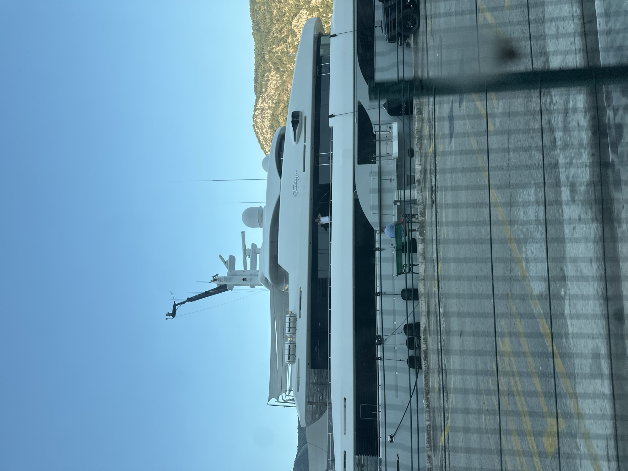 A large, white superyacht named "Taliga" is docked in the city of Kotor, Montenegro. The yacht is multi-decked and appears to be luxurious. In the foreground, a green bench sits on a pier, with two individuals seated on it. One person is facing away from the camera, while the other is partially visible. The pier area is paved with yellow markings. Beyond the yacht, a hillside covered in trees rises under a clear blue sky. The overall scene depicts a serene harbor setting.