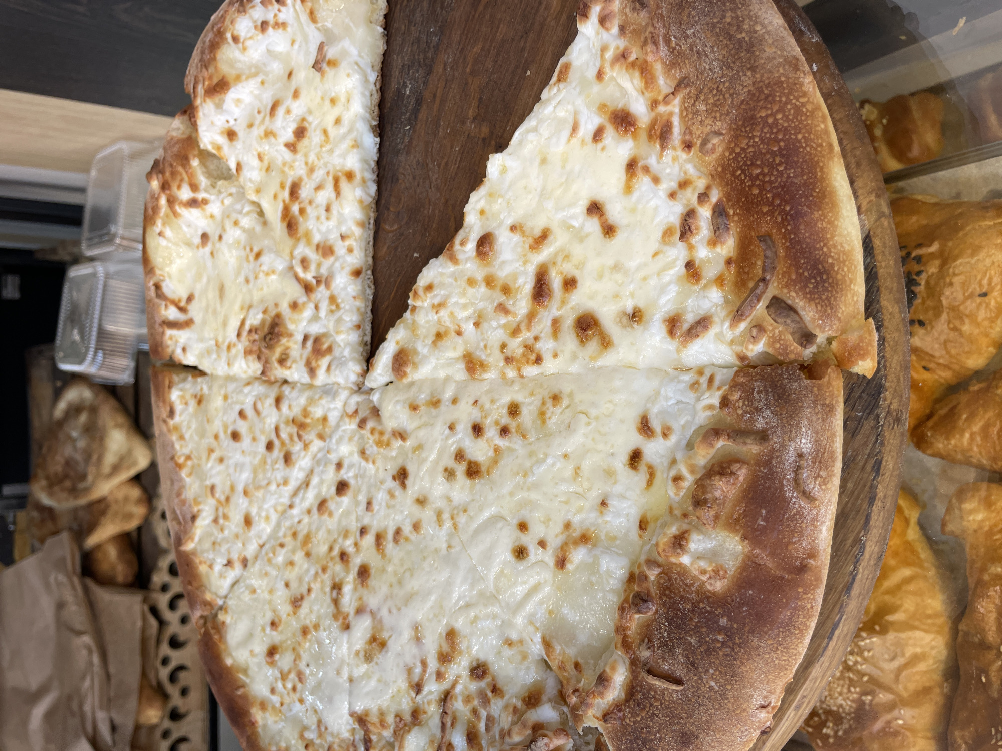 A close-up, overhead shot displays a large, circular cheese pizza cut into four large slices, resting on a wooden board. The pizza has a thick, golden-brown crust with visible bubbles and browning. The surface is generously covered with melted white cheese, showing some areas of golden-brown scorching. In the background, other baked goods and plastic containers are visible, suggesting a bakery or food establishment. The lighting is natural and diffused. The exact location of Sochi, Russia cannot be determined from the image alone.