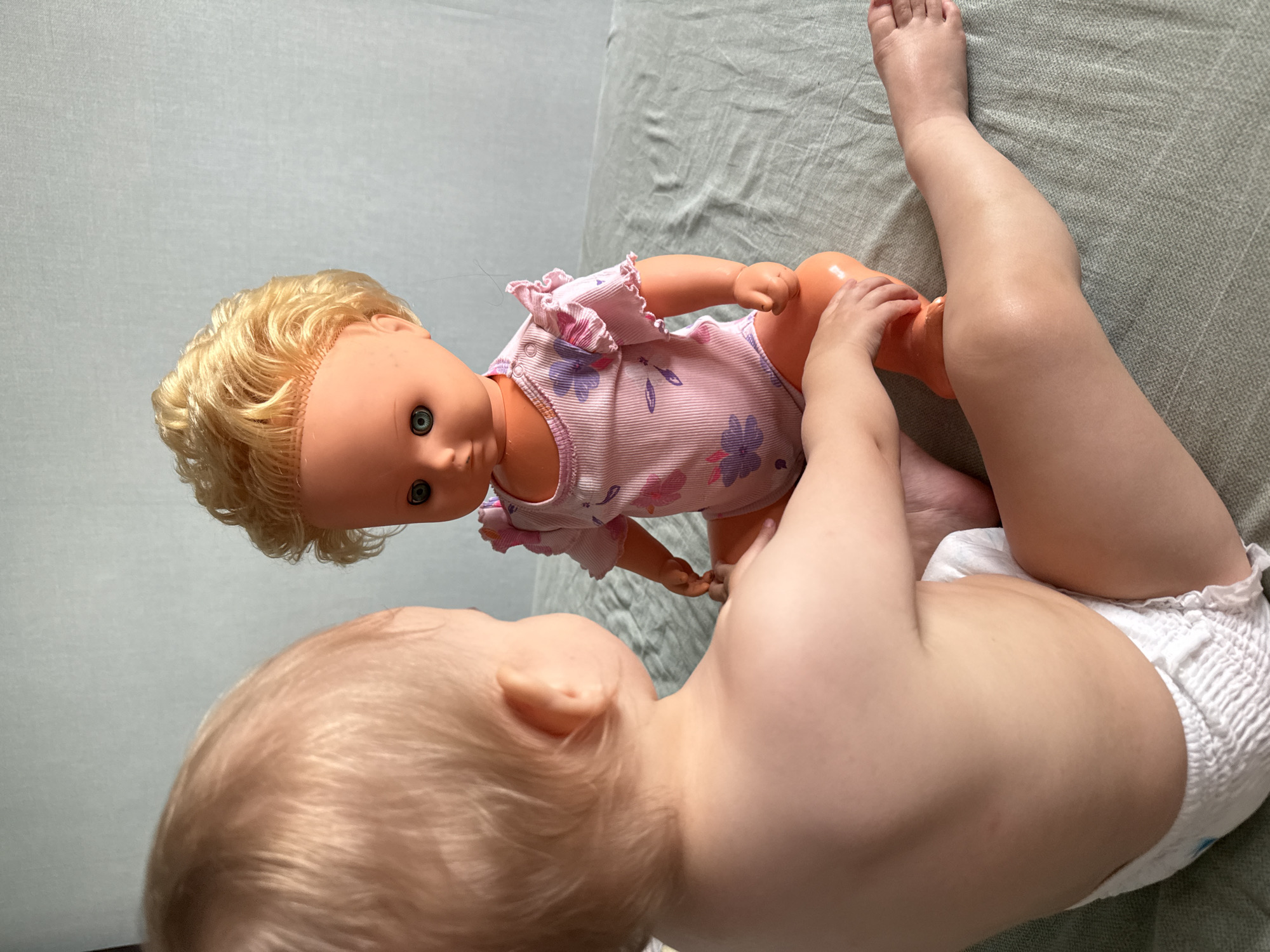 An infant with blonde hair and a diaper is sitting on a textured green surface, facing a blonde-haired doll. The doll is wearing a pink onesie with a floral pattern and is also seated. The infant's back is towards the camera, and they are looking at the doll. The doll's arms are positioned as if interacting with the infant. The background is a plain, light-colored wall. There is no indication of the image being located in Moscow, Russia.