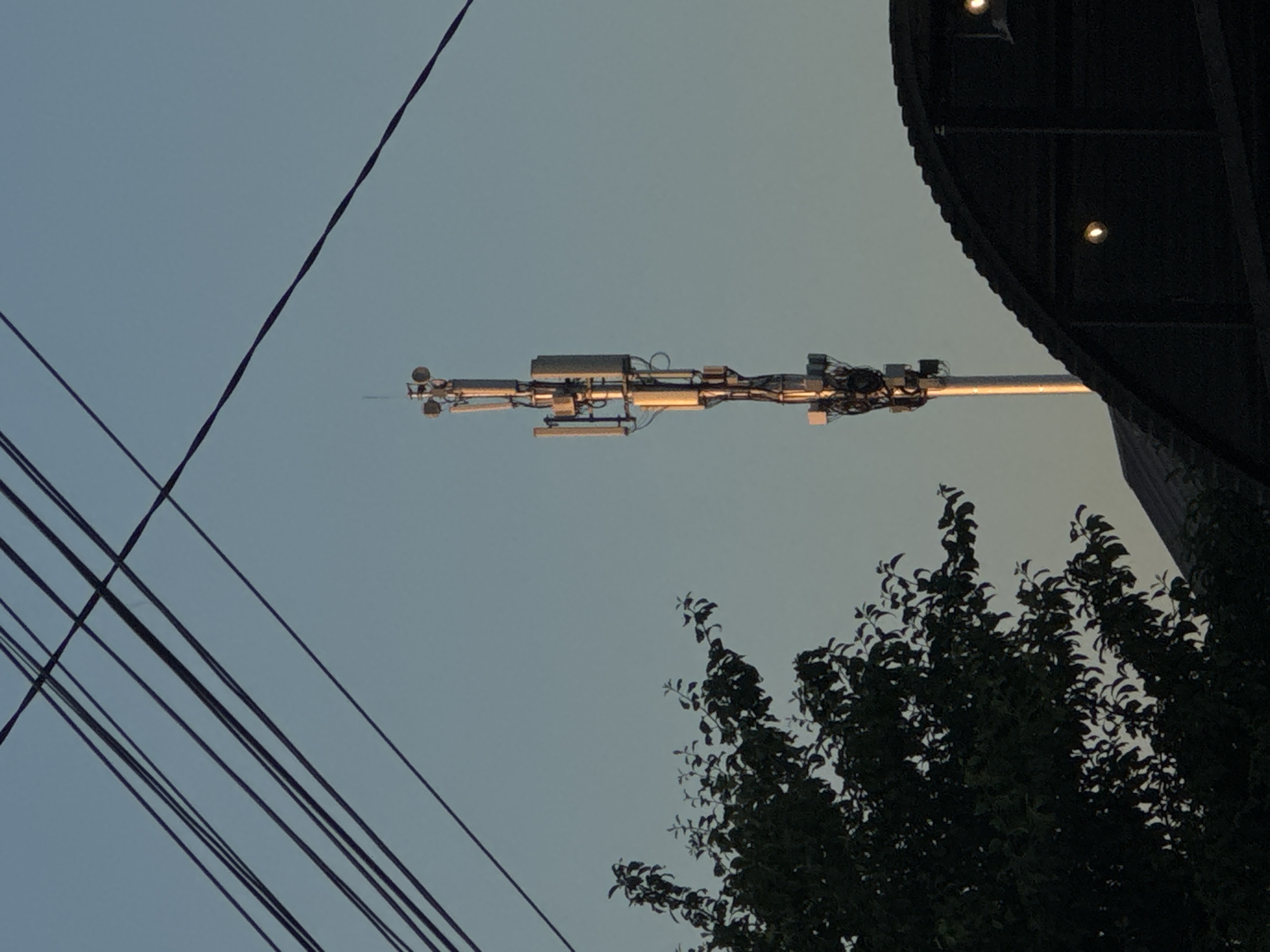 This is a medium-shot photograph taken outdoors at dusk in the city of Mikhaylovsk, Russia. The sky is a clear, pale blue, with several dark power lines diagonally crossing the upper portion of the frame. A tall, slender cell tower, illuminated by the setting sun, stands prominently in the mid-ground, to the right of center. The tower is equipped with multiple antennas and various electrical components. To the left of the cell tower, a dark, leafy tree is partially visible. In the foreground, the dark, curved roof of a building with visible lights obstructs the lower right quadrant of the image. There are no people or animals in the frame.