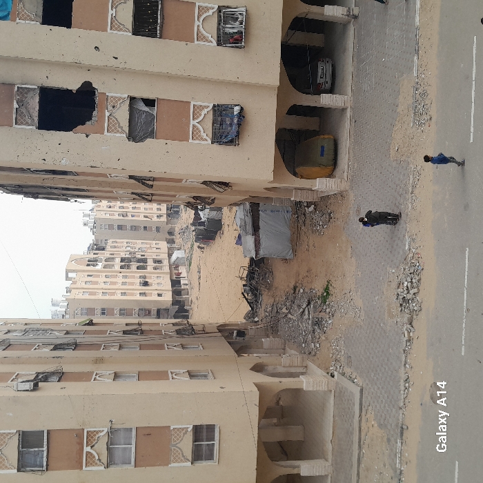 Residential buildings stand on either side of a street with debris scattered along the edges. Tents and makeshift structures are visible amidst the rubble. Some buildings show signs of damage, with broken windows and structural issues. Two people are walking on the street. A car is parked in a shadowed area. The photograph is taken from an elevated perspective, presenting a view of the urban landscape.