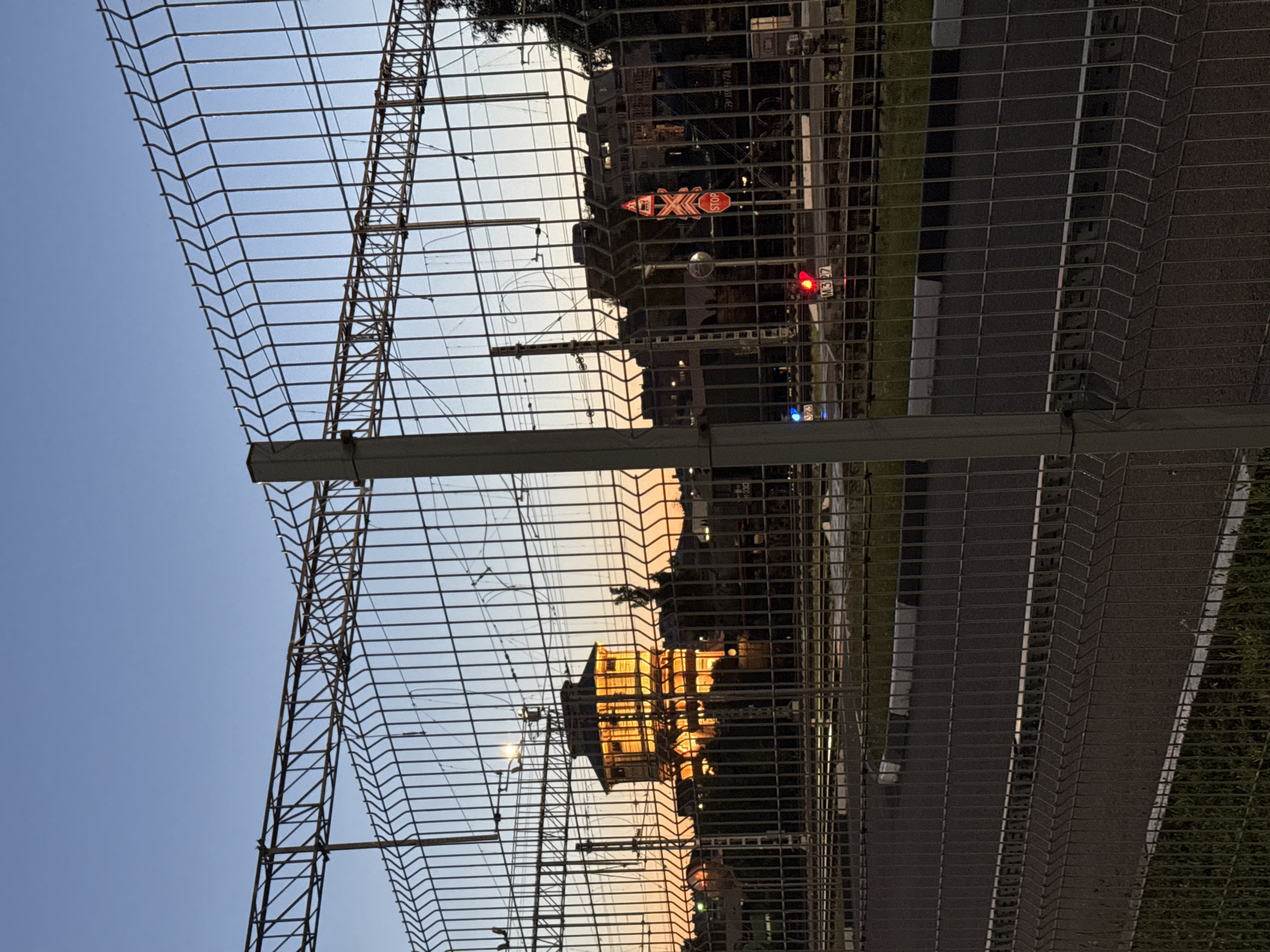 The image captures a view through a metal fence at dusk in Moscow, Russia. In the background, a distinctive wooden building, illuminated with warm yellow light, stands out against the twilight sky. The building appears to be a historic structure, possibly a water tower, given its design. Train tracks and overhead power lines are visible, indicating the location is near a railway. A stop sign is prominently displayed on a traffic signal, and a train signal light shows red. The foreground features a paved road with white and gray markings. The overall scene suggests a transitional period between day and night, with artificial lights beginning to illuminate the urban landscape.