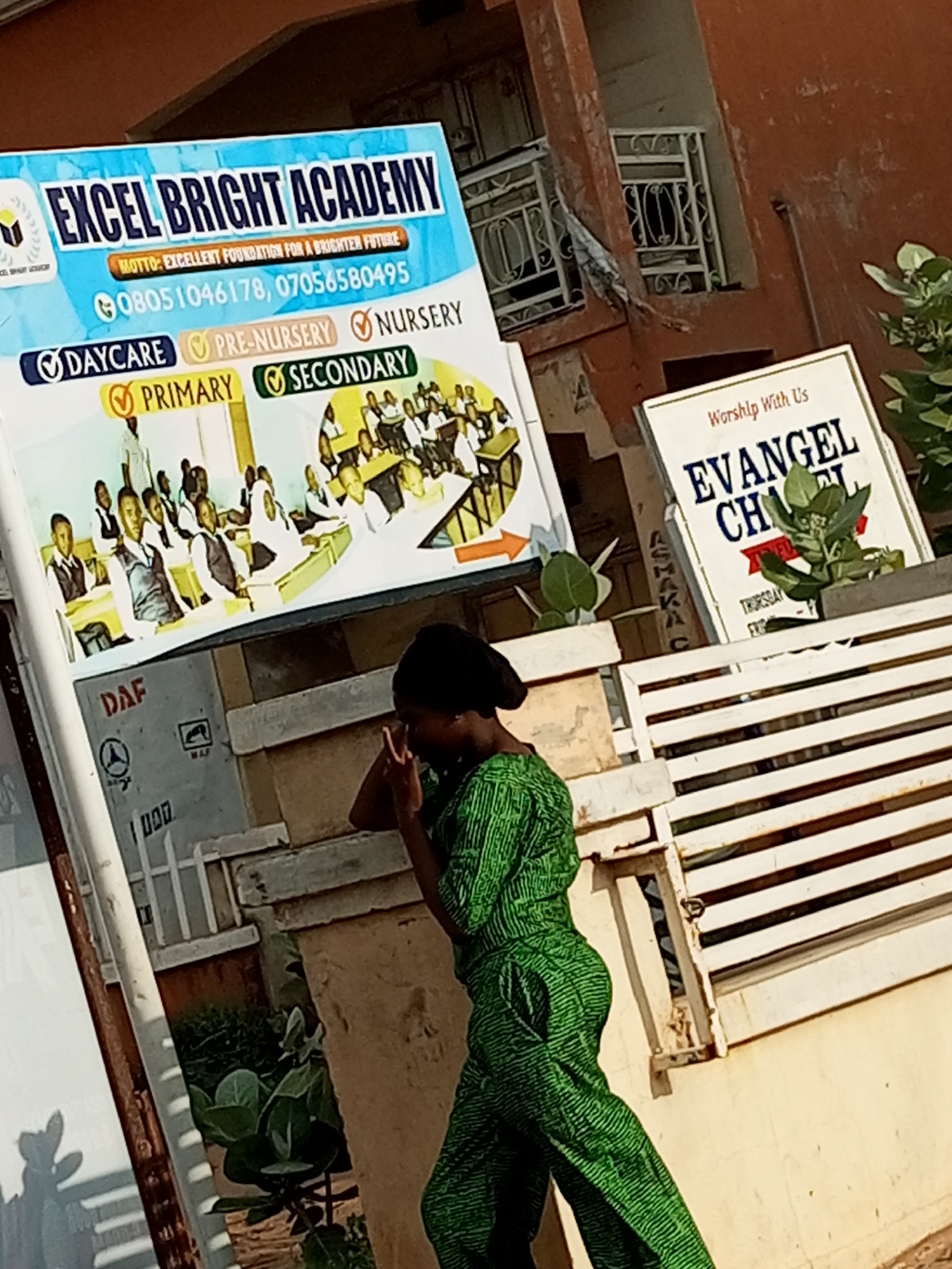 In Tumfure, Nigeria, an outdoor scene captures a woman in a vibrant green patterned outfit, walking past a sign for "Excel Bright Academy." The academy sign, with a blue background, prominently displays the school's name in large white letters, followed by its motto: "Excellent Foundation for a Brighter Future." Below this, contact information is listed as "08051046178, 07056580495." Educational levels offered are highlighted with checkboxes: "Daycare," "Pre-Nursery," "Primary," and "Secondary." A stylized illustration of students in a classroom setting adorns the bottom portion of the academy sign.
To the right of the academy sign, another sign with a white background advertises "Worship With Us EVANGEL CHURCH." A small green plant emerges from a planter in front of this church sign, partially obscuring the word "CHURCH." The overall setting appears to be a street or public area, with hints of buildings and a light-colored bench visible in the background.