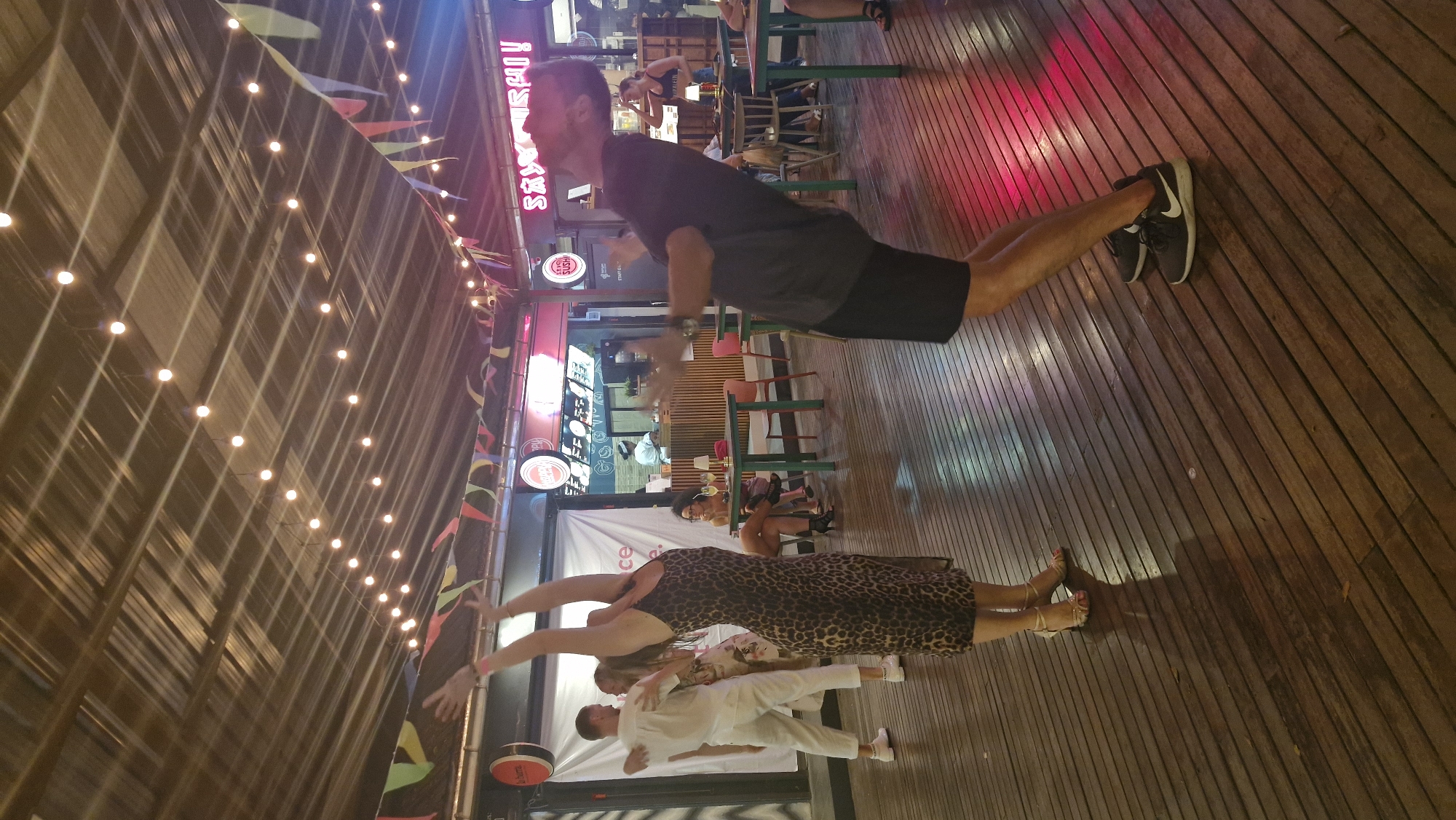 The image depicts an outdoor dining and entertainment area, possibly a restaurant or bar, with a wooden floor and a slatted roof adorned with string lights and colorful pennant banners. In the foreground, a man in a dark t-shirt and shorts is captured mid-dance, leaning backward with his arms extended. To his left, a woman in a leopard-print dress is also dancing, with her arms raised. Behind them, another couple dressed in white appears to be dancing or posing. Further in the background, patrons are seated at tables, some of which are set with drinks. A counter or bar area with food displays is visible, and neon signage, including one that reads "SAUSURISHI!", suggests the establishment offers sushi. The overall atmosphere suggests a lively evening setting. The exact location within Ban Bang Rak, Thailand, is not specified in the image itself.