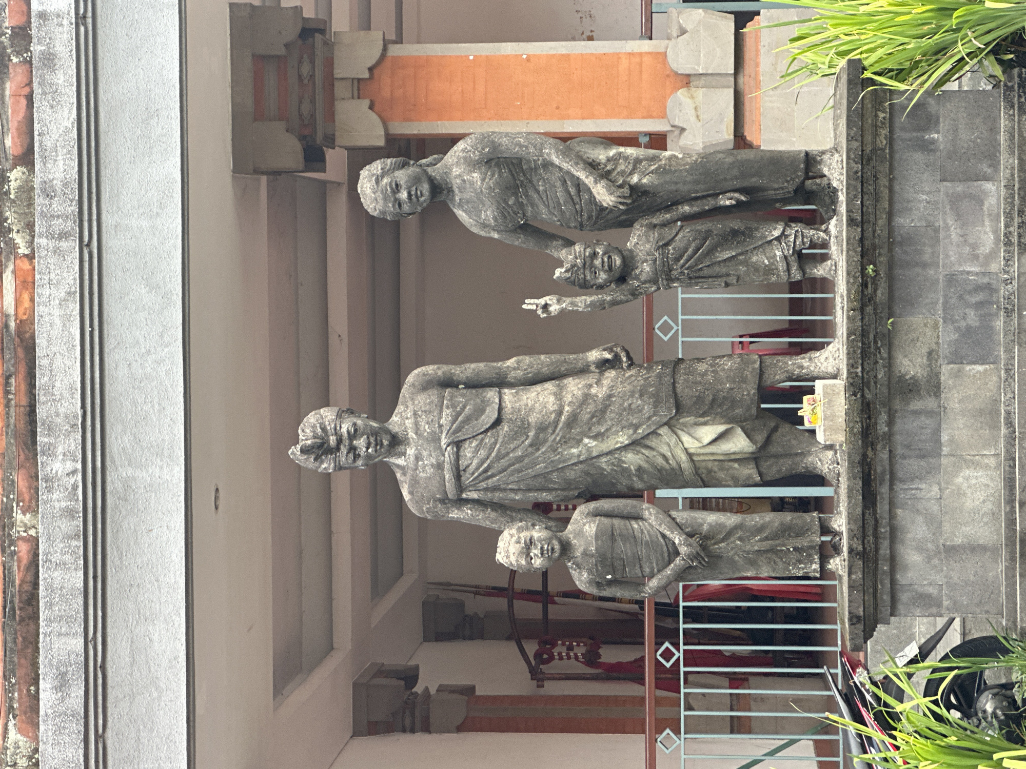 This is a medium-shot, eye-level, outdoor photograph of a stone statue of a family of four in Ubud District, Indonesia. The statue depicts a man and woman standing behind two children, a boy and a girl. The family is dressed in traditional Balinese attire. The man wears a sarong and a headdress. The woman wears a sarong and a top. The boy wears a sarong and a headdress, and he is raising one hand in the air, making a peace sign. The girl wears a sarong. The statues are standing on a stone pedestal in front of a building with orange and white pillars and a red tiled roof. There is a blue metal railing in front of the statues. A motorcycle is parked to the left of the statue, and green plants are growing in front of the pedestal.
