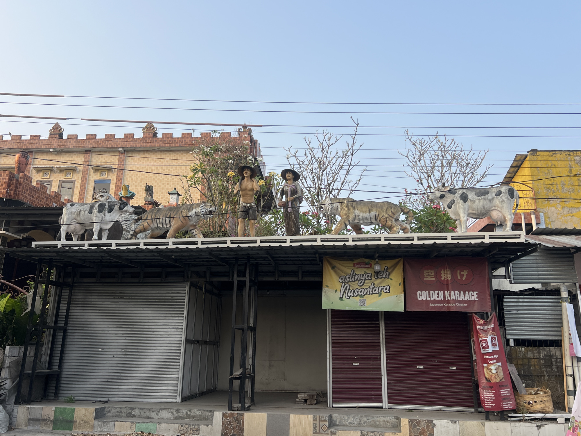 This is an eye-level, medium shot of a storefront in Tlogoadi, Indonesia. The storefront appears closed, with metal roll-up doors. Above the storefront, a decorative roof holds several statues. These include two life-sized statues of cows, two life-sized statues of tigers, and two human-like statues wearing hats and traditional clothing. A yellow banner with green lettering hangs on the storefront. To the right of the yellow banner, a red banner with Japanese and English text reads "GOLDEN KARAAGE Japanese Karaage Chicken". The sky above is clear and light blue.