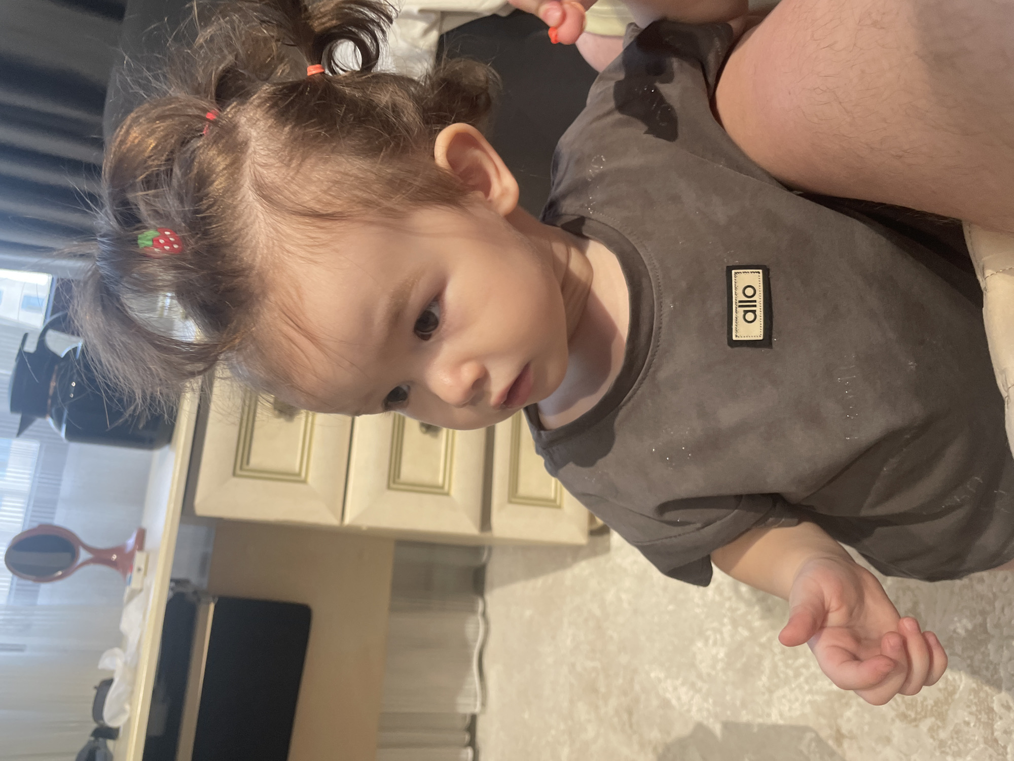 A toddler with brown hair pulled into messy pigtails, adorned with strawberry-shaped hair clips, sits indoors. The child wears a dark gray t-shirt with a rectangular black patch bearing the word "allo" in white letters. The child is looking to the left. The background shows a dresser with light-colored drawers and a blurry white curtain. The floor is covered with a light-colored textured rug. The scene appears to be a domestic setting within Dushanbe, Tajikistan.