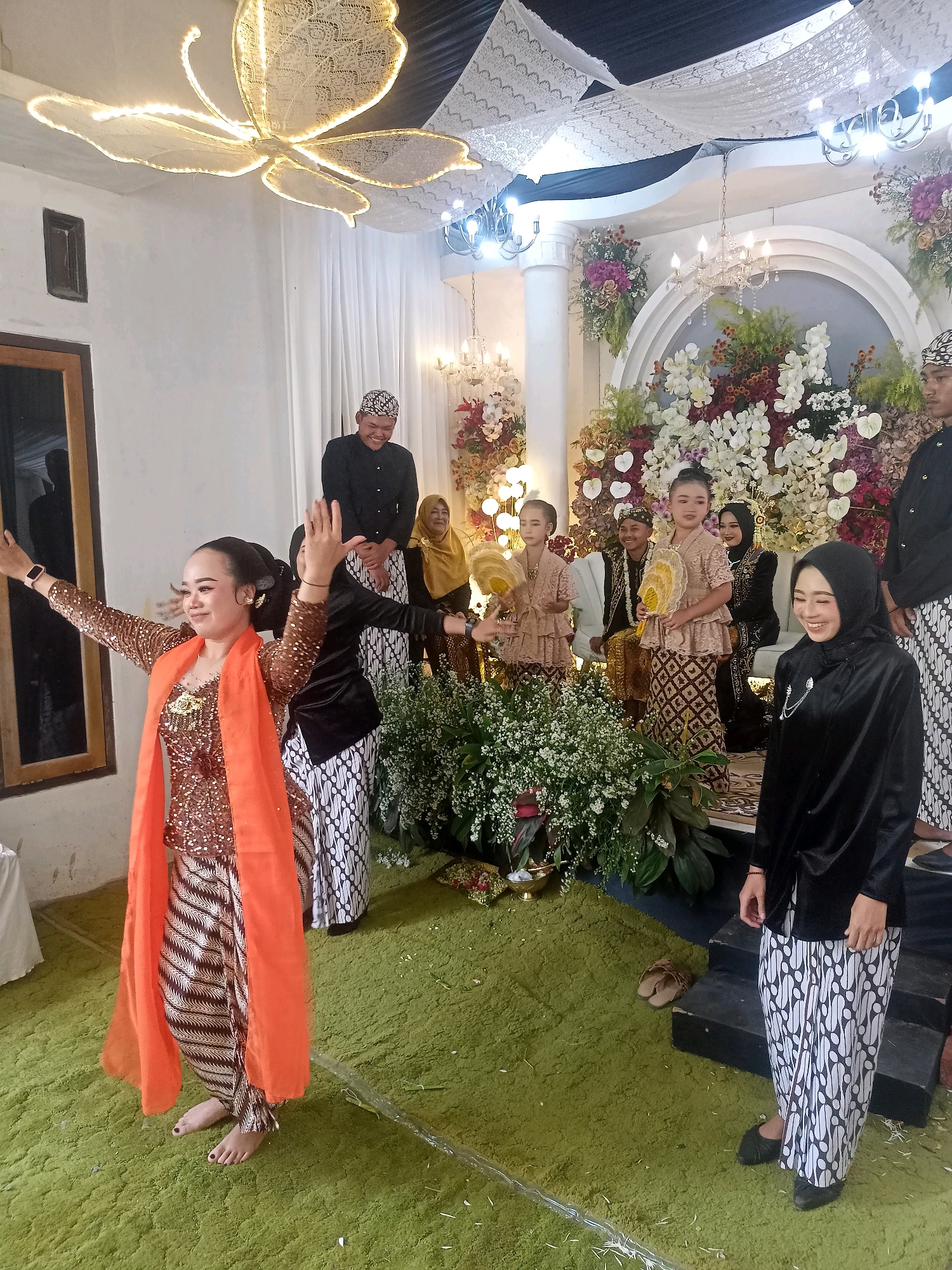 The image depicts a cultural event, possibly a wedding, taking place indoors in Badamita, Indonesia. The room is elaborately decorated with white lace draped from the ceiling and numerous floral arrangements, featuring white, pink, and deep red flowers, along with lush green foliage. A large, butterfly-shaped light fixture illuminates the space.
In the foreground, a woman is captured in mid-dance, wearing a shimmering bronze and gold sequined top and a traditional batik sarong. She has an orange sash draped around her shoulders and is barefoot, with painted toenails. To her right, another woman in a black headscarf and black attire, also wearing a batik sarong, smiles warmly.
Behind the dancing woman, several individuals are present. A man in a black shirt and batik sarong stands tall, smiling. A group of children and adults are seated or standing near a floral backdrop, some holding decorative fans. One young girl is dressed in a formal, light-colored traditional outfit, and another woman wears a golden headscarf. Another man, in a black shirt and batik sarong, stands slightly further back.
The floor is covered in a thick, bright green carpet, with some dried white petals scattered on it, indicating a recent ceremonial event. A reflective plastic sheet or covering is visible on the carpet in the lower part of the image. The overall ambiance suggests a festive and traditional celebration.