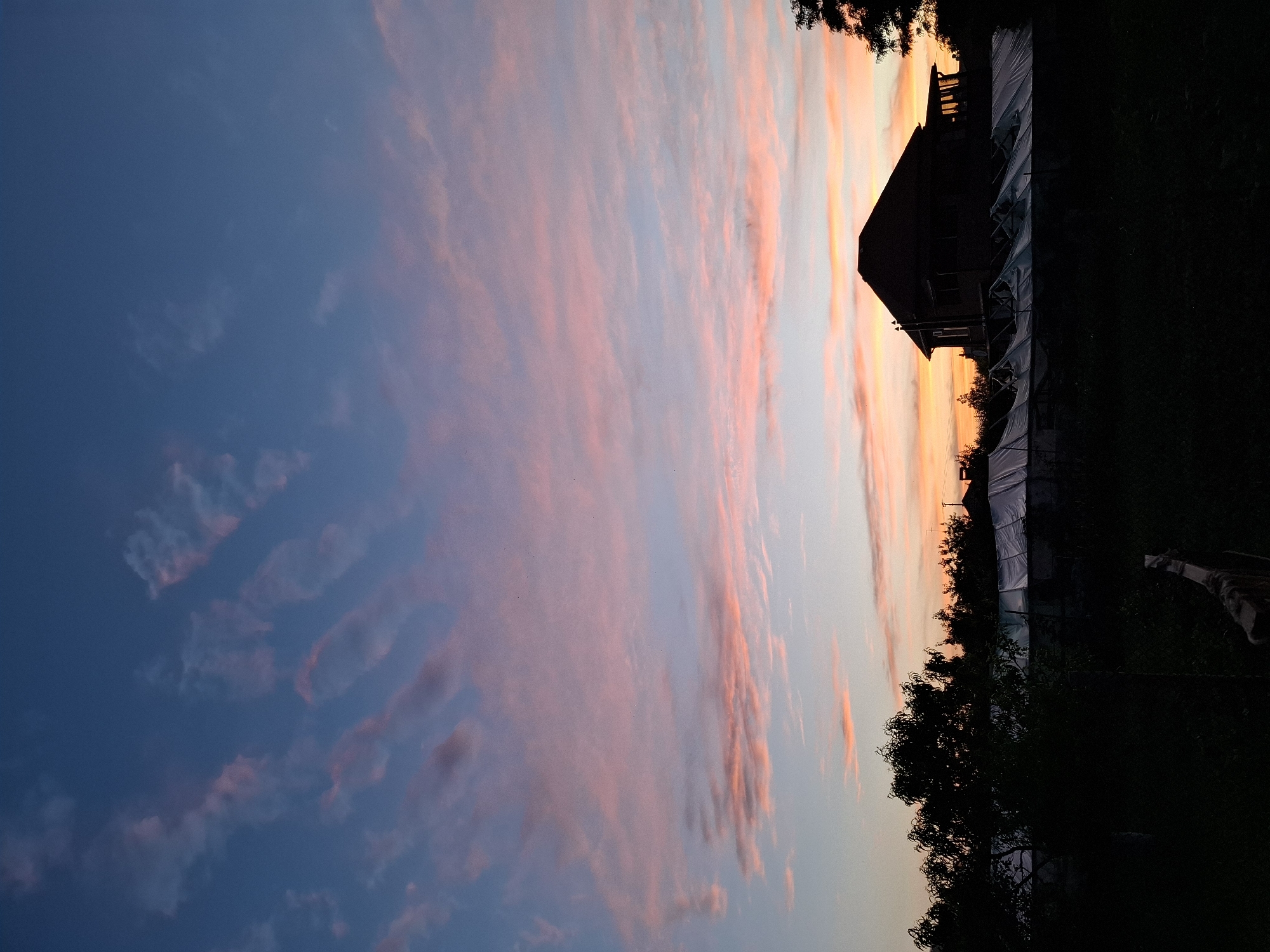 The image captures a twilight scene with a vibrant sky filled with streaks of pink and orange clouds against a deep blue backdrop. In the foreground, silhouetted against the fading light, is a residential building with a prominent roof and a covered structure resembling a greenhouse or extension. Several trees, also in silhouette, frame the scene from the left and right sides. The ground in the foreground is dark and indistinct, suggesting it is either nightfall or deeply shaded. The image appears to be taken from a residential area, potentially a backyard or garden, with the sky dominating the upper half of the frame. The location is specified as Korenovsk, Russia.
