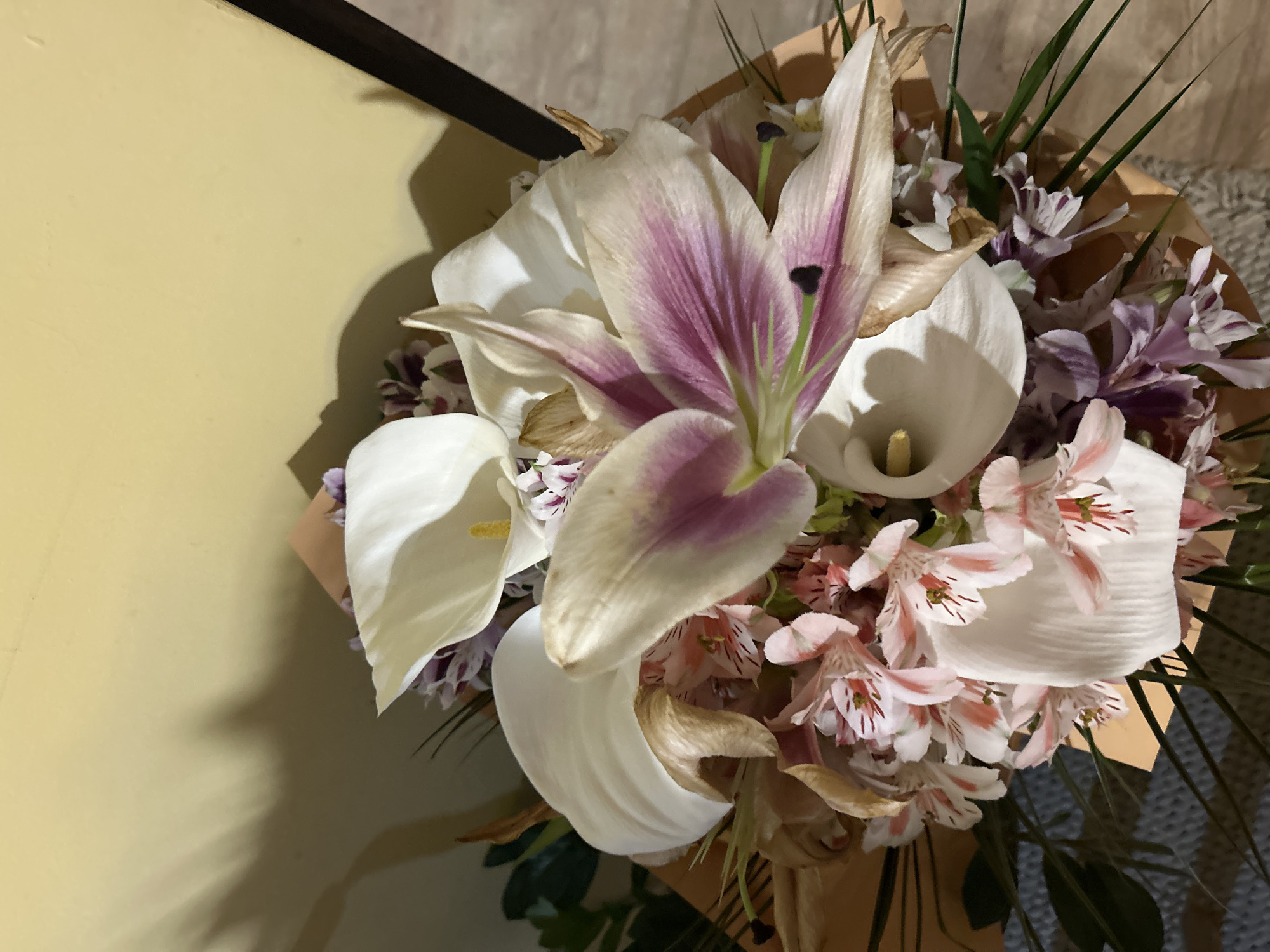 A close-up shot of a bouquet of flowers is displayed against a muted yellow wall and a floor covered in a grey rug. The bouquet consists of predominantly white calla lilies, one prominent lily with pink-purple streaks, and pink and purple striped Alstroemeria. The flowers are bundled in light brown wrapping. Some of the flowers appear slightly wilted, while others are fresh. Thin green stems are interspersed throughout the bouquet. The scene is indoors. The location, Burgas, Bulgaria, is inferred and is not directly evidenced by the image.
